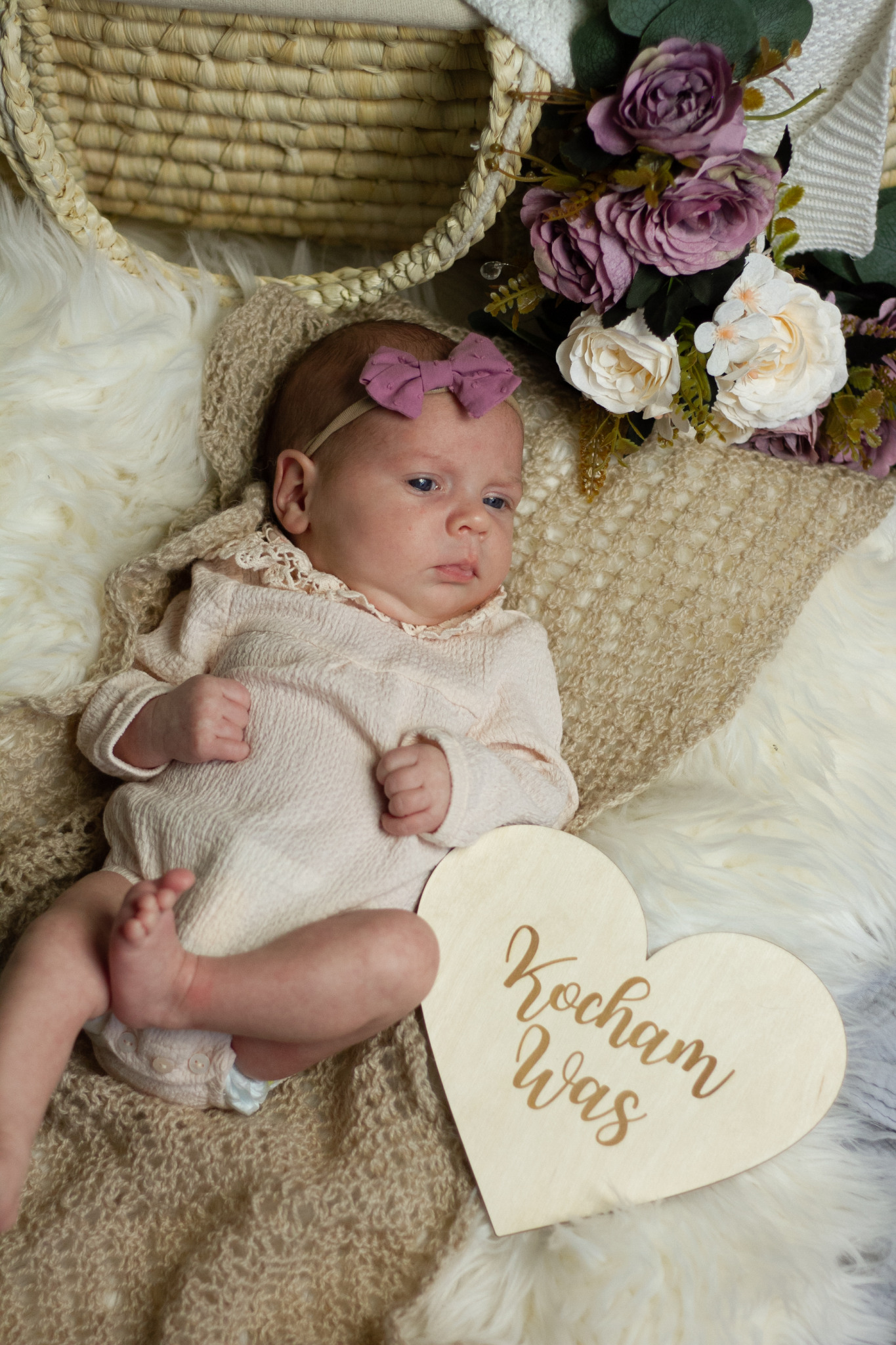 Noworodek w beżowym ubranku i fioletową kokardką na głowie leży na futrzanym kocyku obok drewnianego serca z napisem 'Kocham Was', kwiaty i koszyk w tle.