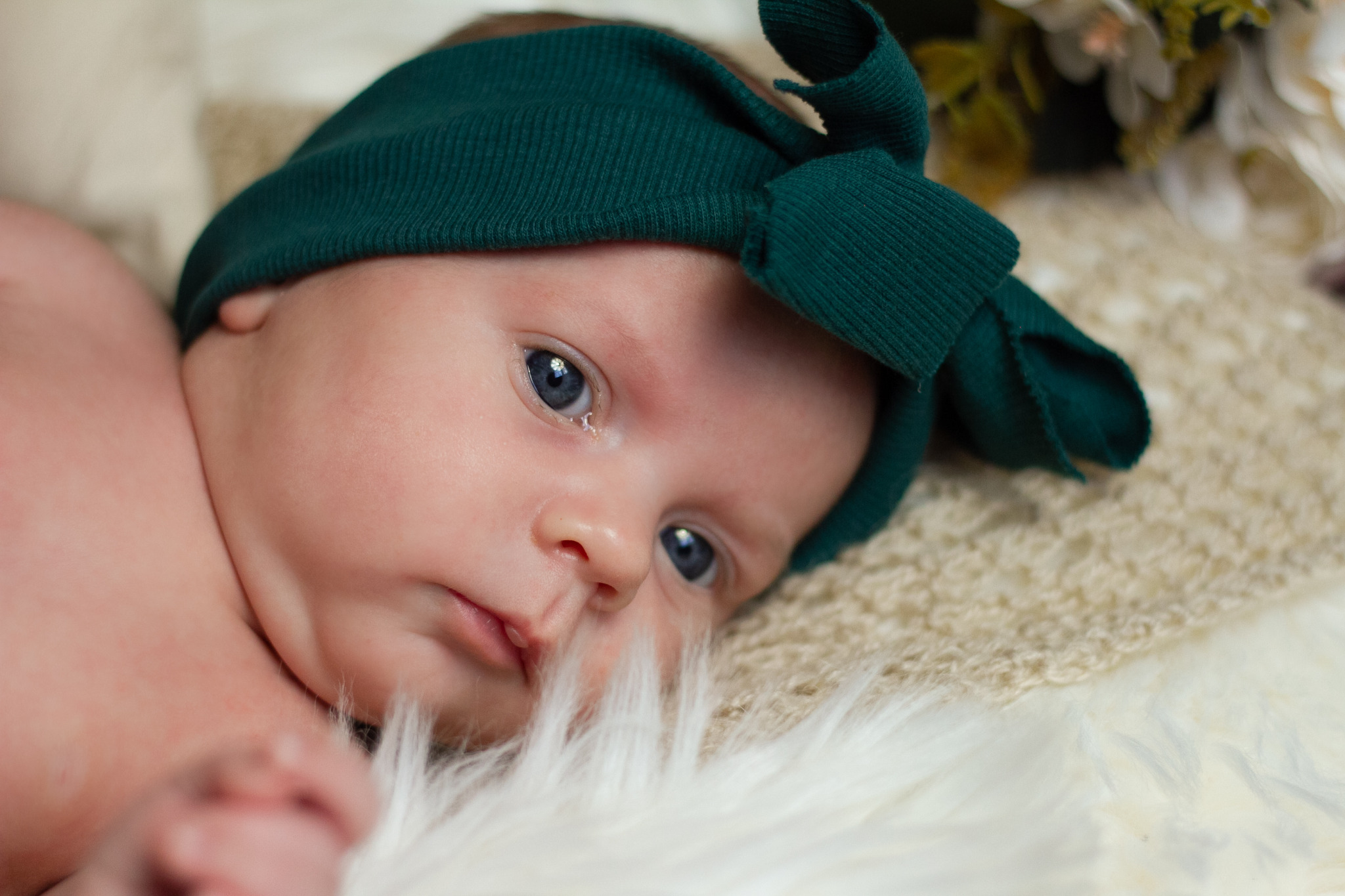 Portret noworodka z dużymi, niebieskimi oczami i zieloną opaską na głowie, leżącego na kremowym i białym futerkowym tle.