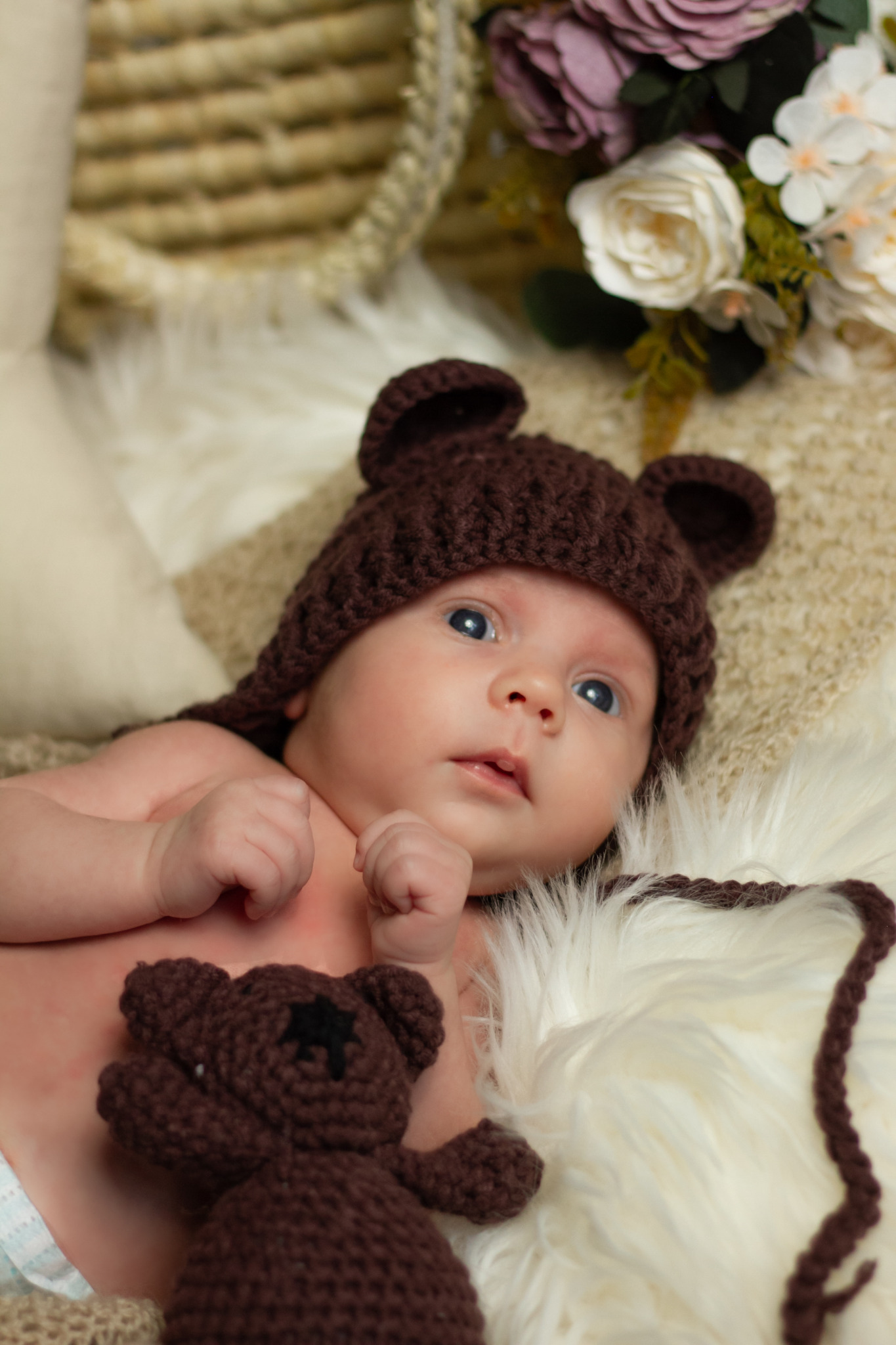 Noworodek leżący na białym futrze, ubrany w brązową czapkę z uszami misia, obok pluszowy miś, w tle kosz z kwiatami.
