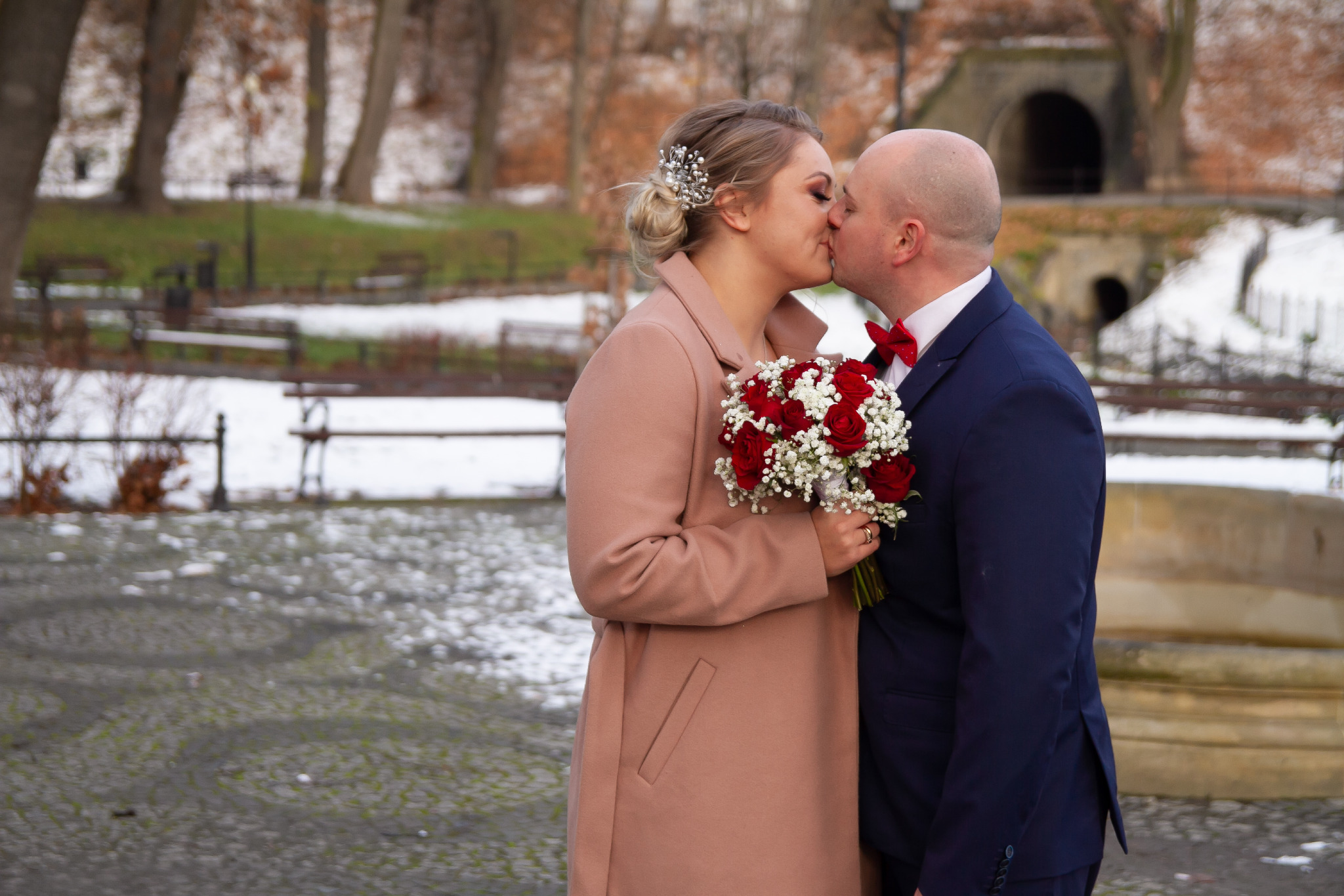 Para młoda całuje się na tle zimowego parku z fragmentami śniegu, panna młoda w płaszczu w kolorze pudrowego różu trzyma bukiet czerwonych róż i białych gipsówek, a pan młody w granatowym...