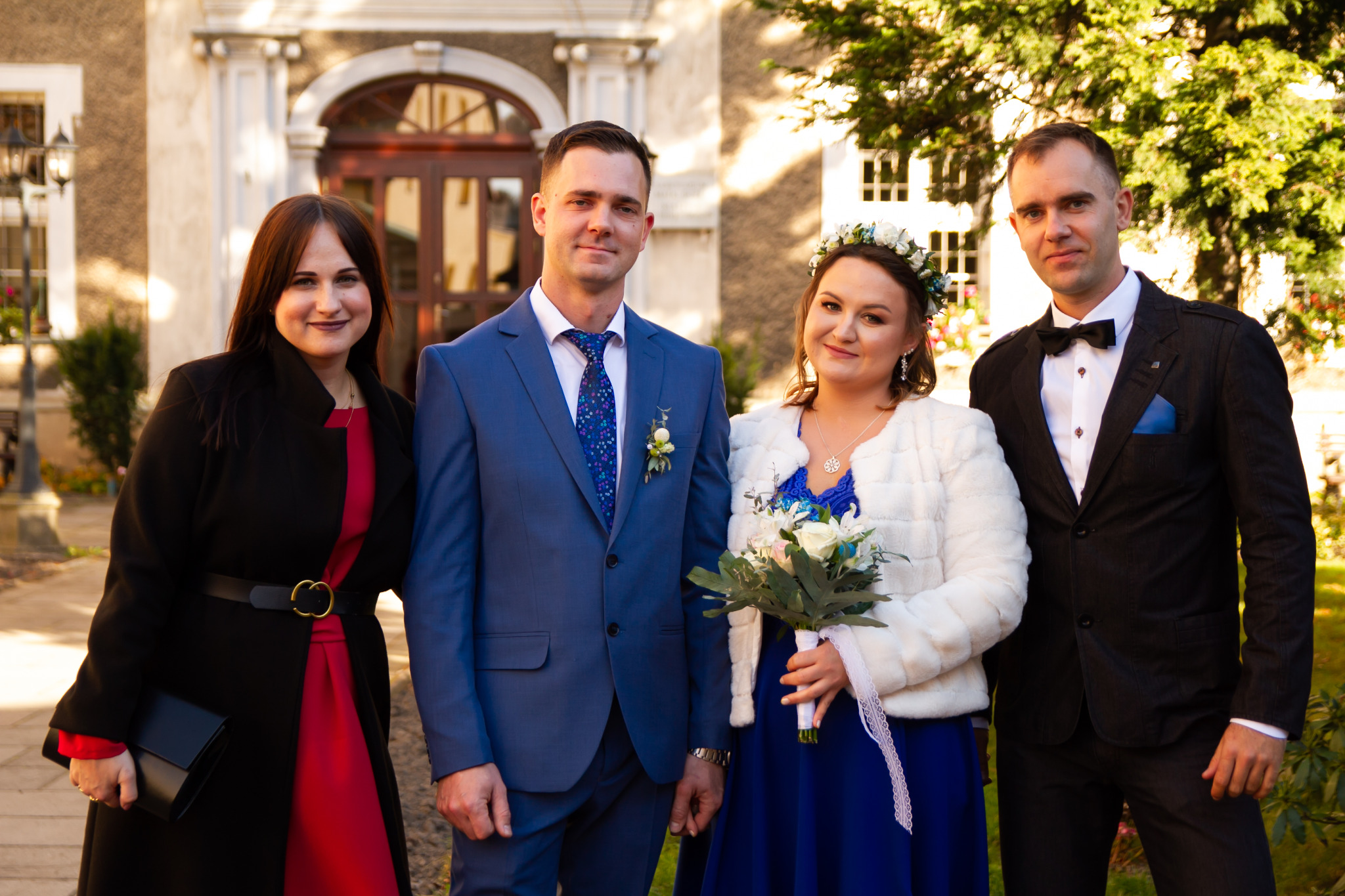 Uśmiechnięta para młoda w towarzystwie elegancko ubranych gości pozuje na tle budynku z łukowatym wejściem i zieleni. Panna młoda w białym wianku i futerku trzyma bukiet, pan młody w niebieskim...