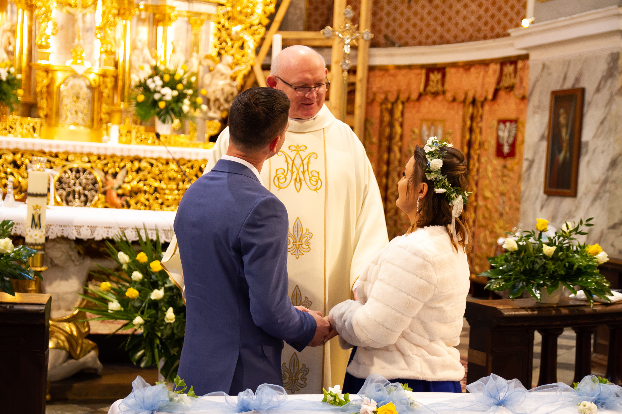 Para młoda trzymająca się za ręce podczas ceremonii ślubnej w bogato zdobionym kościele, z księdzem w tle i kwiatowymi dekoracjami.