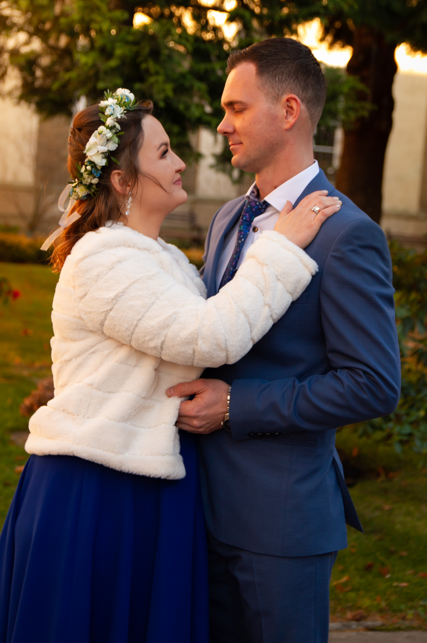 Para młoda w objęciach, ona w sukni z kobaltowym dołem i wiankiem z białych kwiatów na głowie, on w niebieskim garniturze i wzorzystym krawacie, patrzą na siebie z czułością na tle zieleni i budynku.