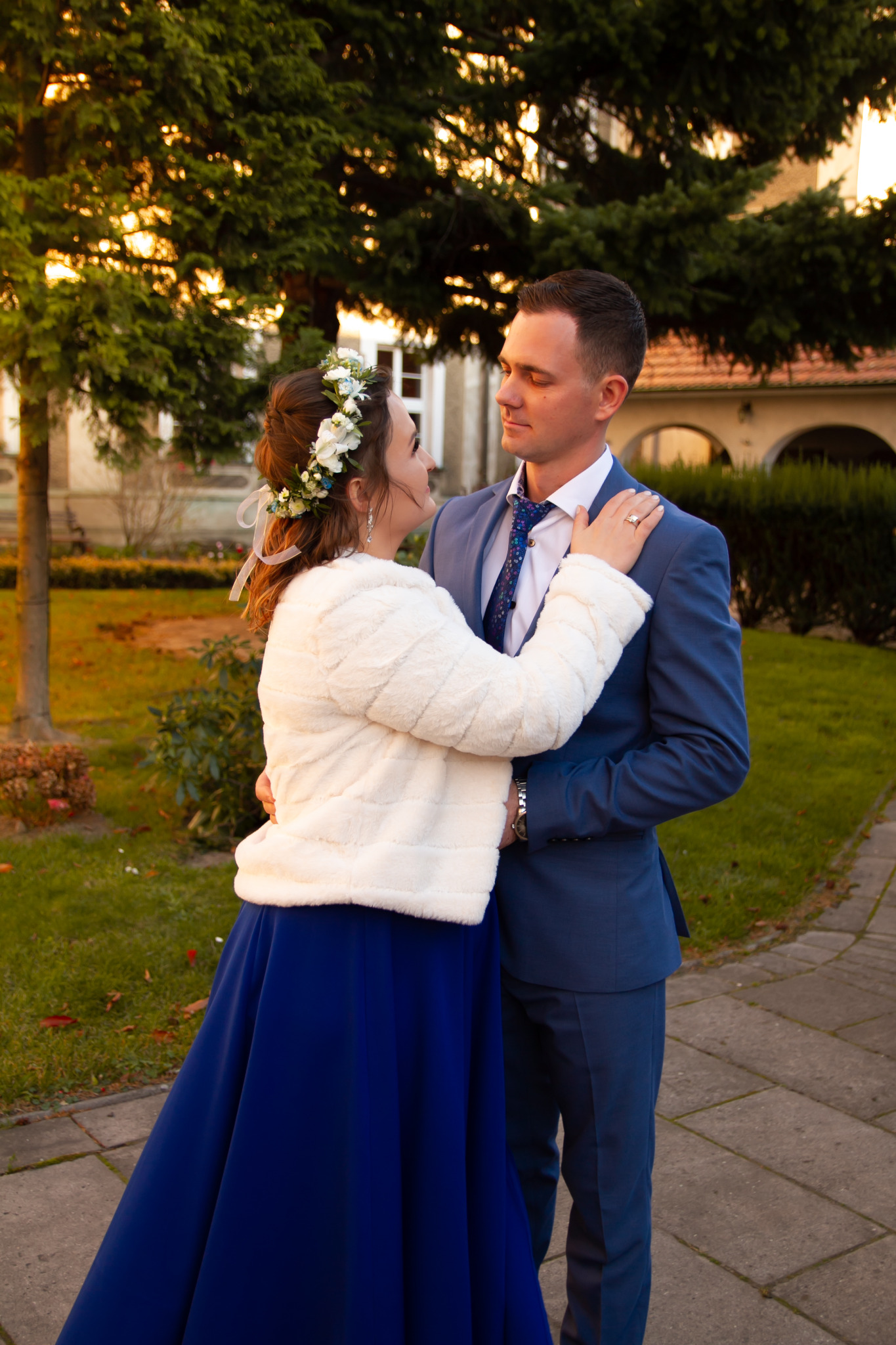 Para młoda obejmująca się na tle drzew i budynku w ciepłym świetle zachodzącego słońca, ona w białym futerku i sukni w kolorze kobaltowym, on w niebieskim garniturze.