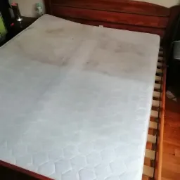 Materac na drewnianym łóżku, widoczne plamy i zabrudzenia na powierzchni.