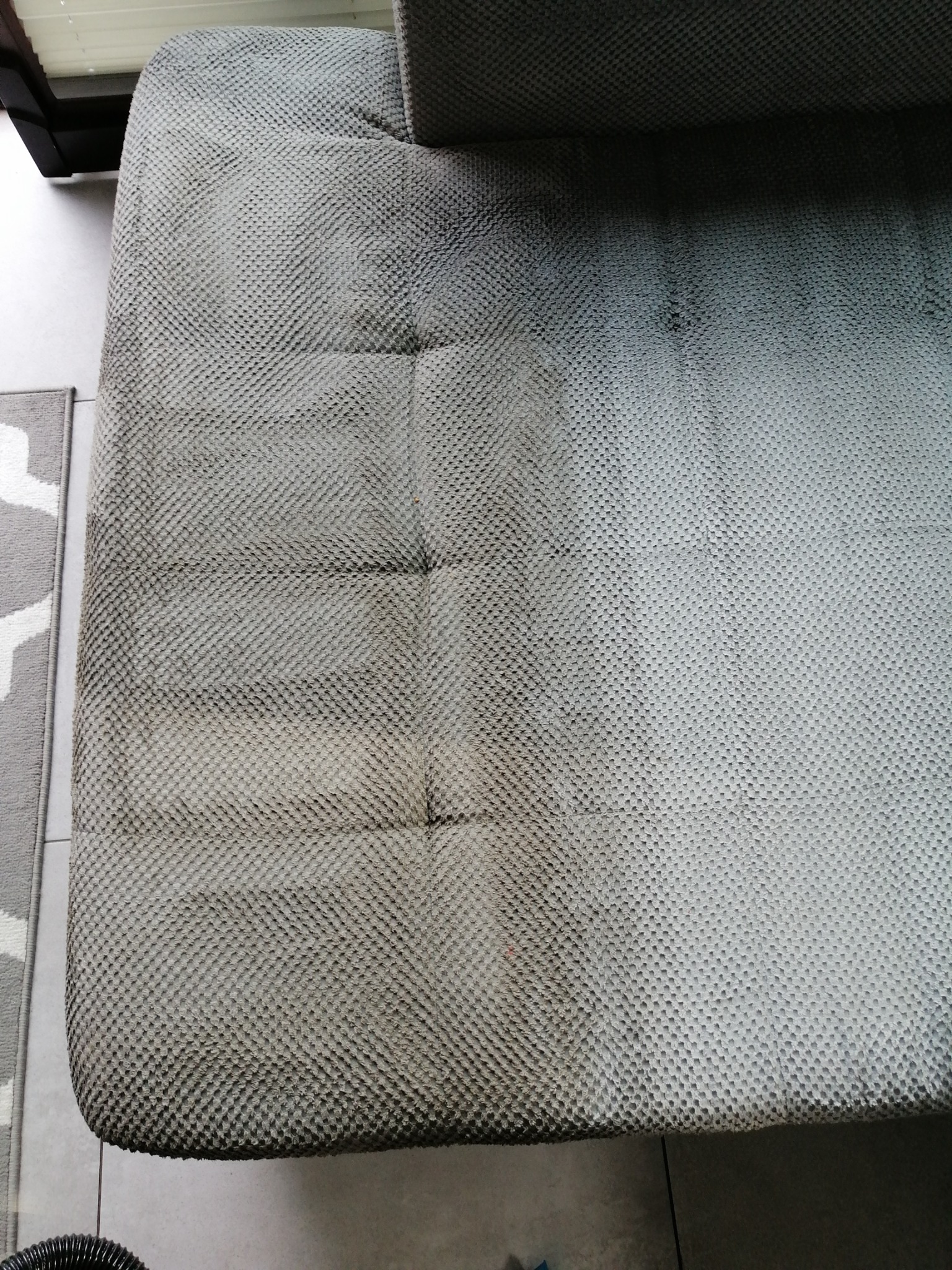 Fragment szarej sofy z wyraźną różnicą w czystości tapicerki: prawa strona świeżo wyczyszczona, lewa strona brudna, widoczna struktura materiału.