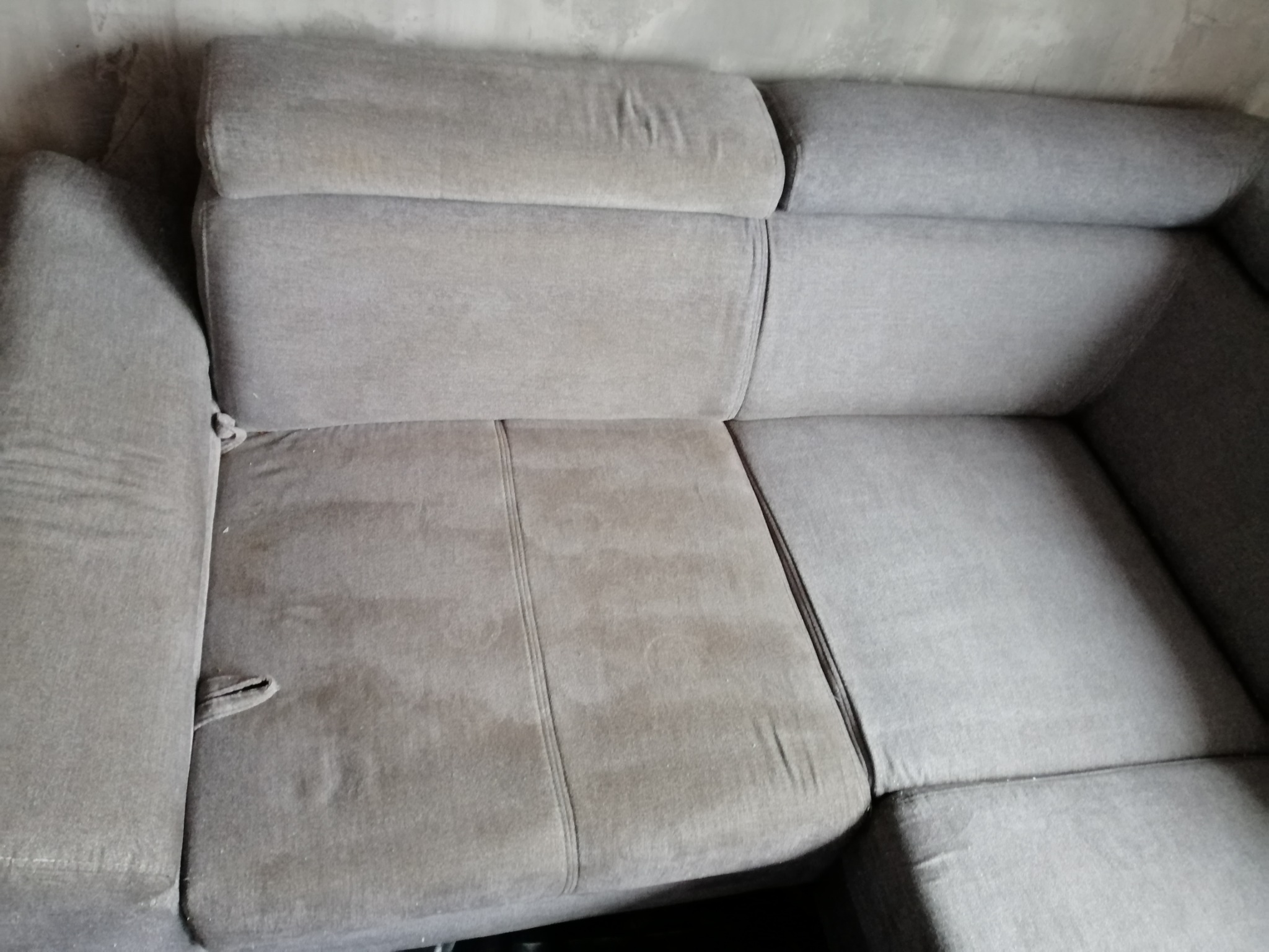 Szara, narożna kanapa z widocznymi plamami na siedzisku, wymagająca odświeżenia tapicerki.