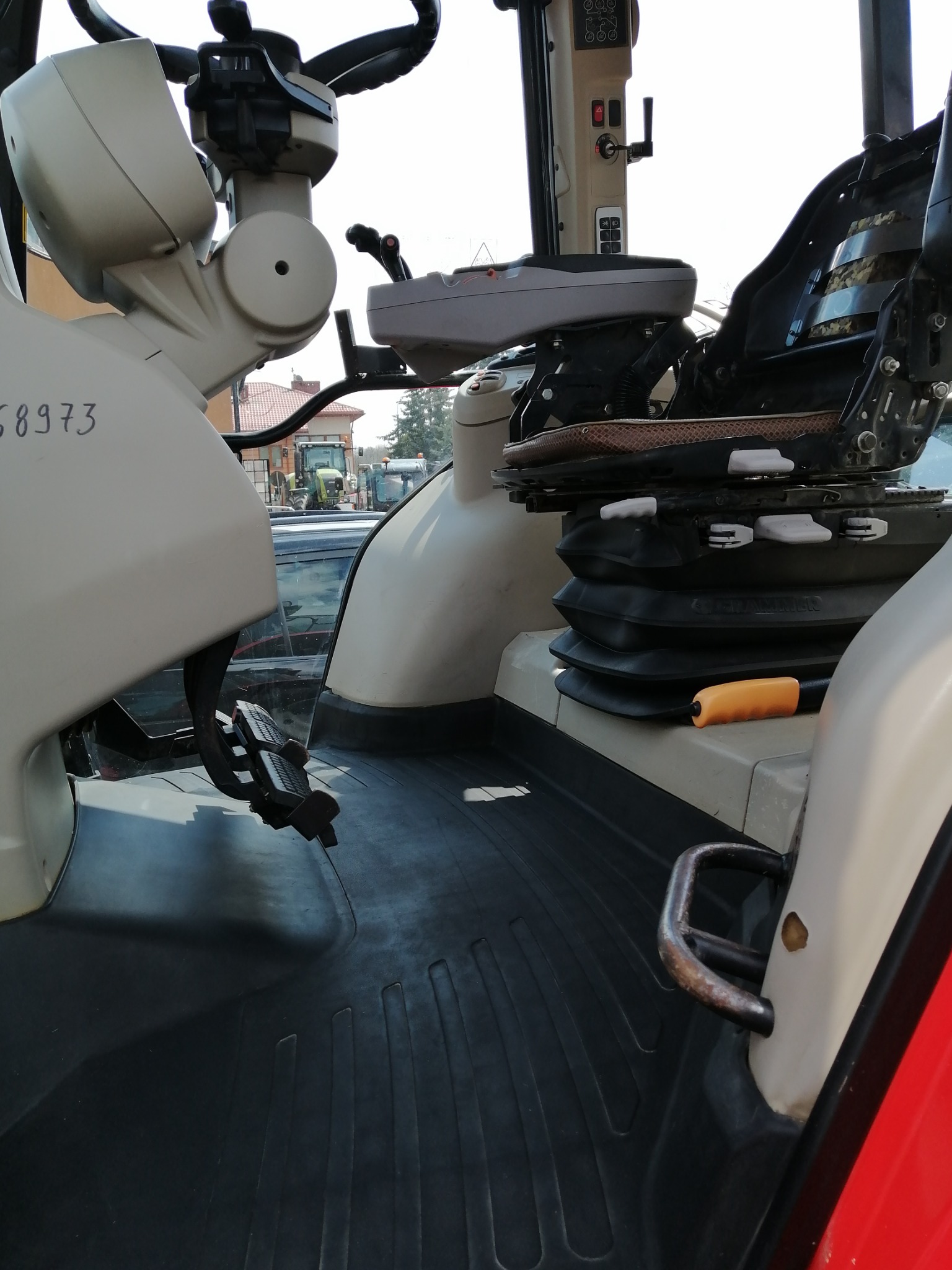 Wnętrze kabiny ciągnika rolniczego, widok na fotel operatora, kierownicę i pedały, z widocznymi elementami sterowania i gumową wycieraczką podłogową.