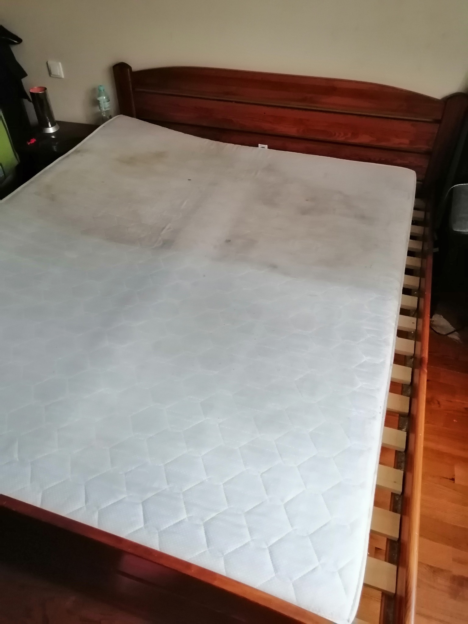 Materac na drewnianym łóżku, widoczne plamy i zabrudzenia na powierzchni.