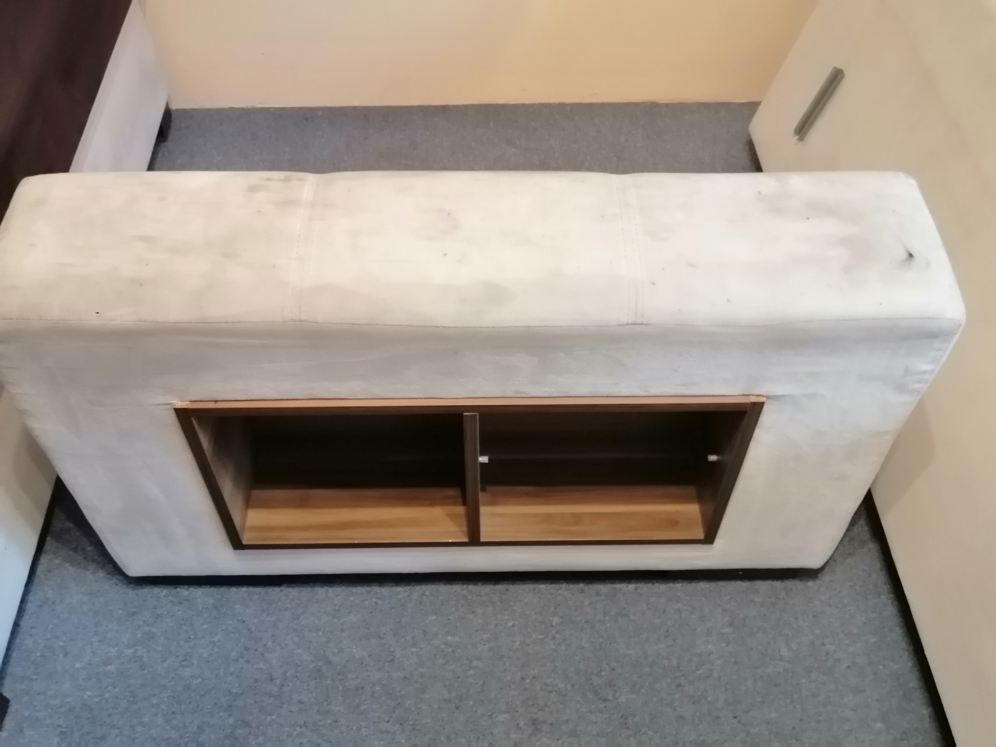 Tapicerowany puf z drewnianą wnęką, widoczne plamy na tapicerce, na szarym dywanie.