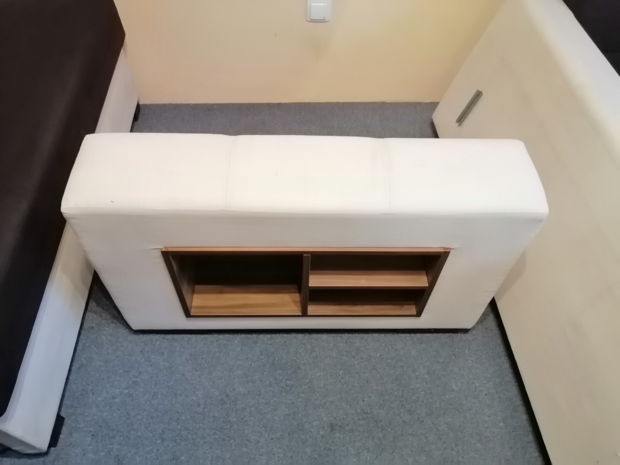 Tapicerowany puf z drewnianymi półkami wkomponowanymi w konstrukcję, stojący na szarej wykładzinie