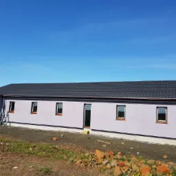 Niski, parterowy budynek z szarą elewacją i ciemną, metalową dachówką, widoczny z zewnątrz na etapie wykończenia, z rozstawioną drabiną i stertą kamieni na pierwszym planie.