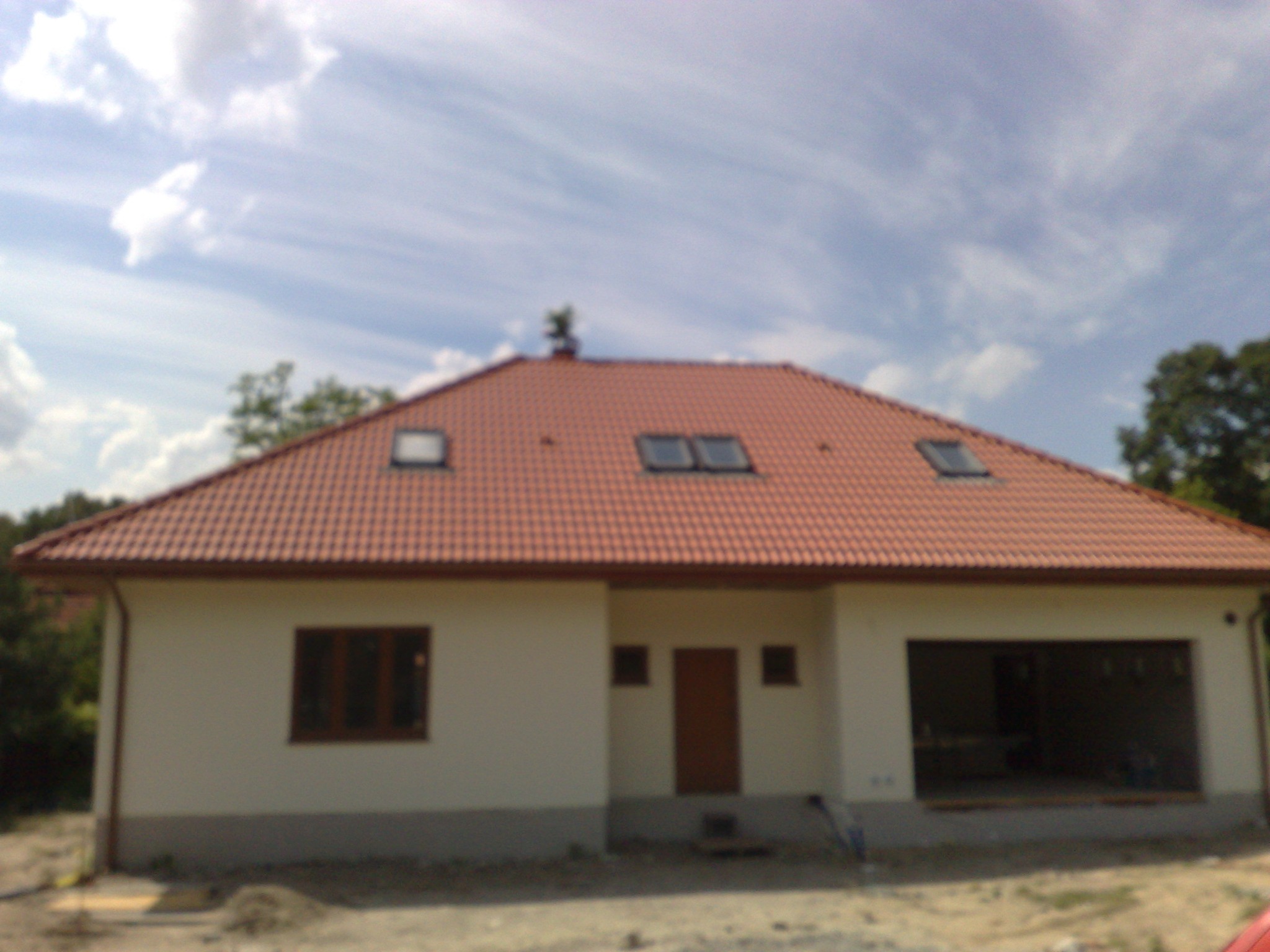 Niedokończony parterowy budynek z czerwoną dachówką, trzema oknami dachowymi i otwartym garażem, widziany w słoneczny dzień.