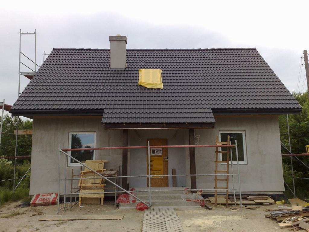 Surowy stan domu z ciemną dachówką, widoczne rusztowania i materiały budowlane, żółta plandeka na dachu, szary tynk elewacyjny.