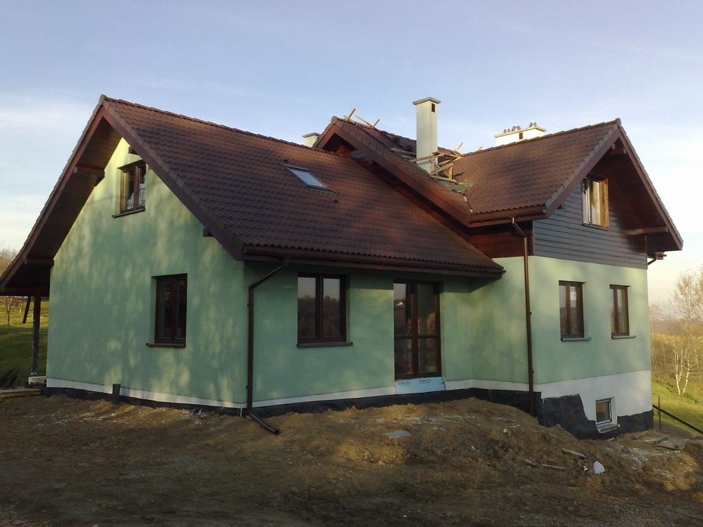 Dwukondygnacyjny budynek z zielonymi ścianami i brązowym dachem, częściowo w trakcie budowy, z widocznymi elementami drewnianej konstrukcji dachu i kominem.