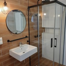 Usługi Rem Bud Grzegorz Misiewicz - Wykończona łazienka we Wrocławiu z drewnopodobnymi płytkami, kabiną prysznicową z czarnymi detalami, umywalką nablatową i okrągłym lustrem z metalową ramą.