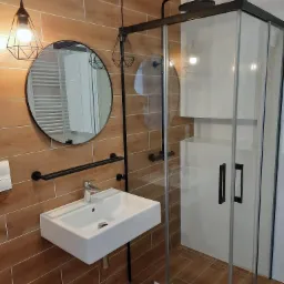 Wykończona łazienka we Wrocławiu z drewnopodobnymi płytkami, kabiną prysznicową z czarnymi detalami, umywalką nablatową i okrągłym lustrem z metalową ramą.