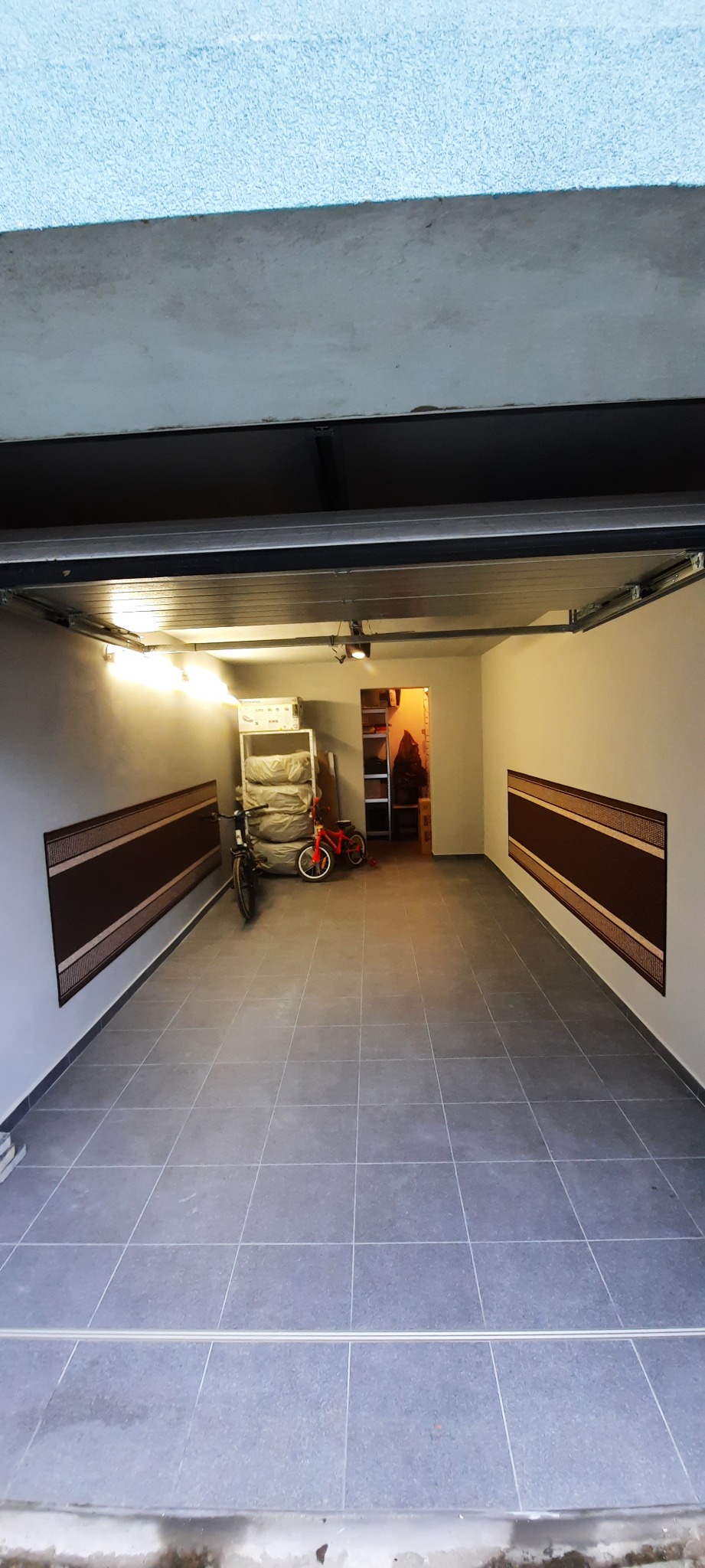 Wyremontowany garaż z szarą, kwadratową podłogą z płytek, białymi ścianami z brązowymi panelami dekoracyjnymi, oświetleniem punktowym i otwartymi drzwiami do pomieszczenia gospodarczego w tle.