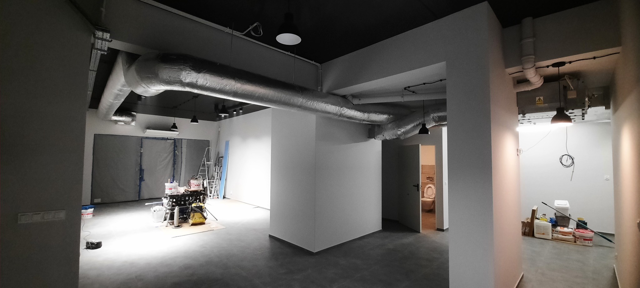 Wnętrze biurowe w trakcie odświeżania, widoczne przewody wentylacyjne pod sufitem, zabezpieczone pomieszczenie folią malarską, drabiny i sprzęt malarski na podłodze, ściany przygotowane do malowania.