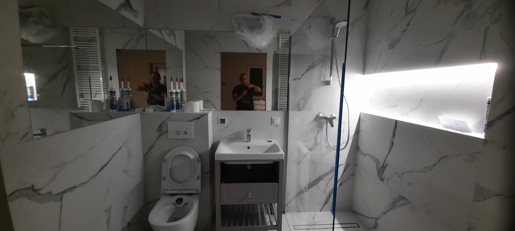 Nowoczesna łazienka z marmurowymi płytkami, kabiną prysznicową typu walk-in i podświetlaną wnęką; widoczne odbicie osoby w lustrze i częściowo zakryte elementy instalacji.