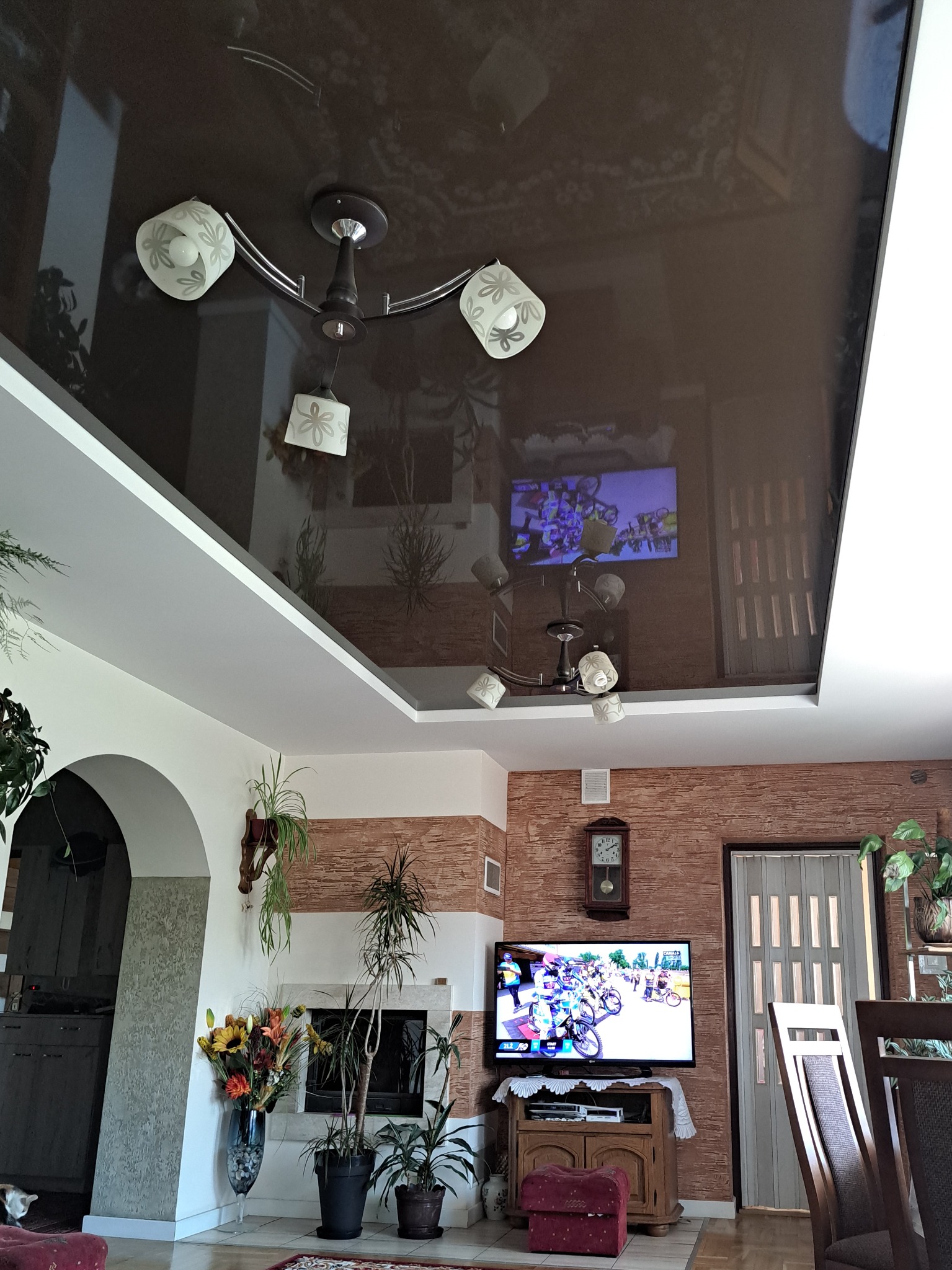 Ciemnobrązowy, błyszczący sufit napinany z odbiciem wnętrza salonu, widoczny żyrandol z białymi kloszami w kwiaty i fragment białego sufitu podwieszanego.