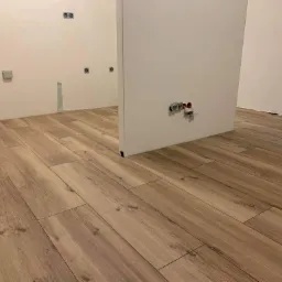 Ułożona podłoga z paneli imitujących drewno, widoczne gniazdka elektryczne i puszki instalacyjne na ścianie w trakcie prac wykończeniowych.