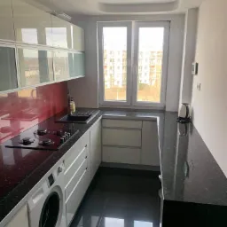 Nowoczesna, jasna kuchnia z czarnym blatem i błyszczącą podłogą, z pralką w zabudowie pod blatem, widok na okno z budynkami w tle.