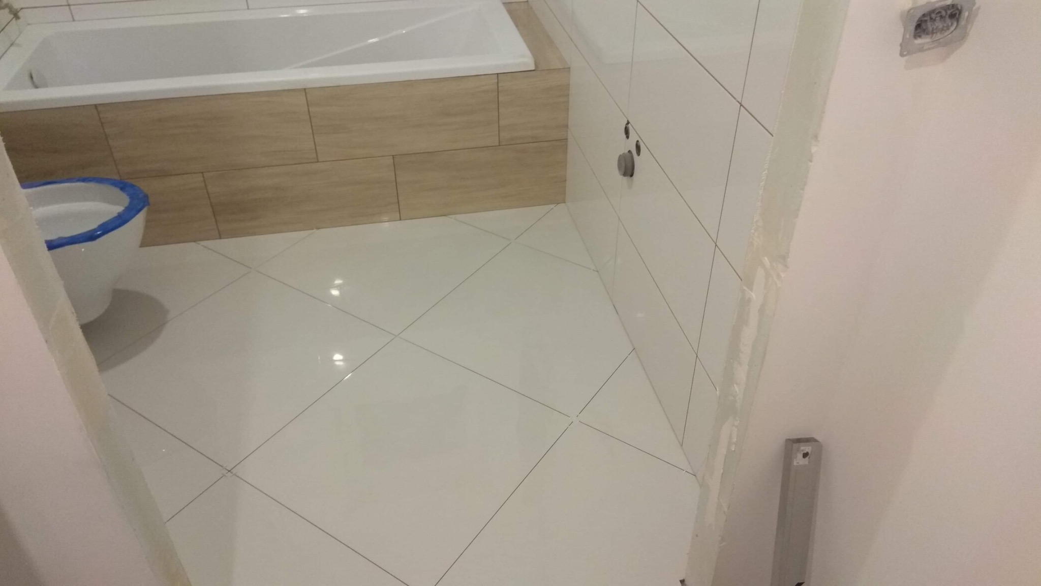 Białe płytki podłogowe ułożone w karo w trakcie prac wykończeniowych łazienki, widoczna wanna obudowana panelami imitującymi drewno oraz zabezpieczona toaleta.