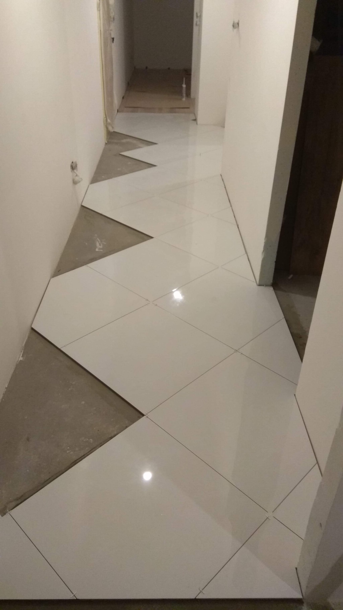 Kładzenie białych, błyszczących płytek podłogowych w korytarzu, widoczne fragmenty surowej wylewki przed fugowaniem.
