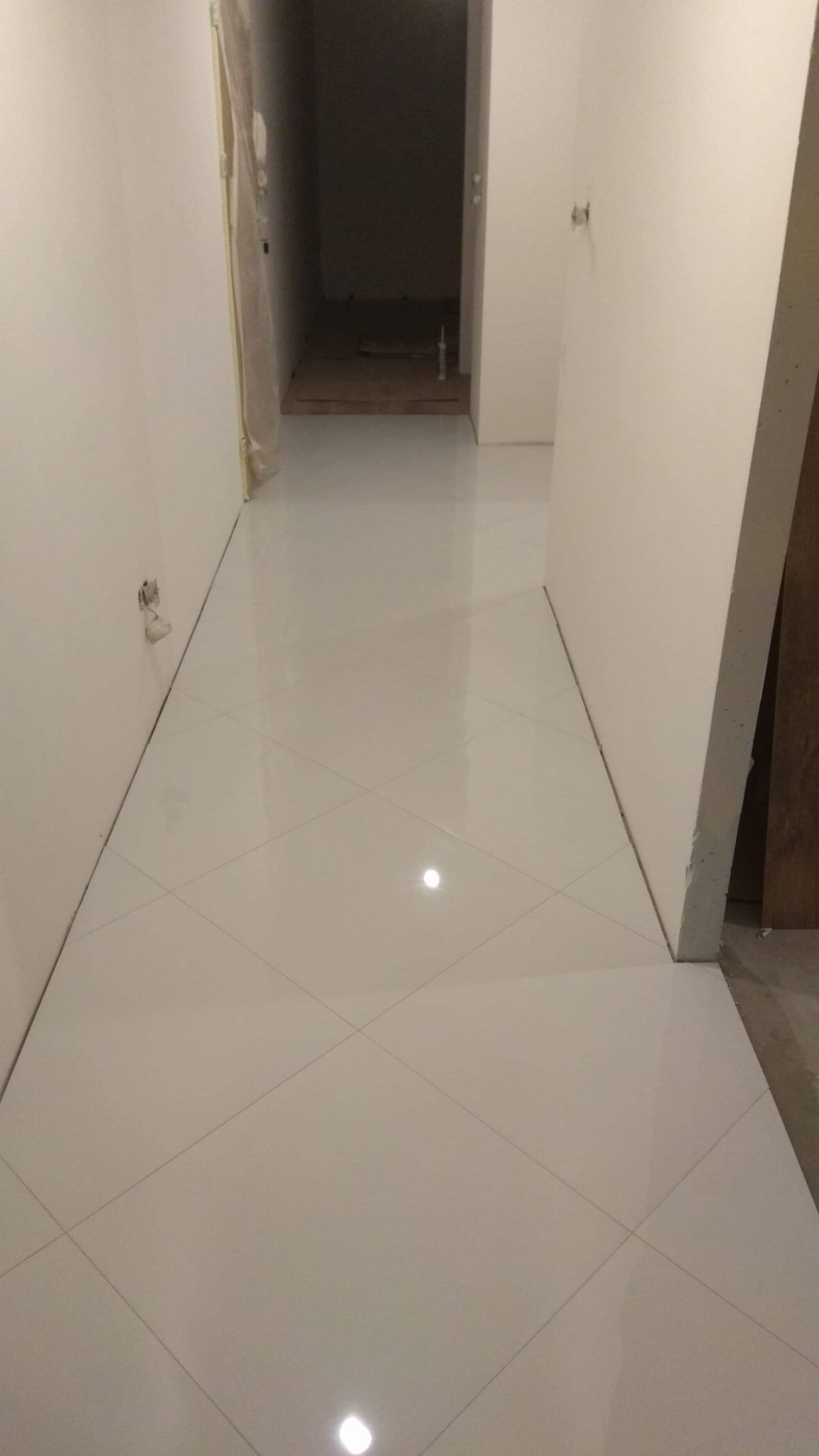 Korytarz z nowo ułożoną, błyszczącą, białą podłogą z płytek ułożonych w karo, widoczne niedokończone ściany i przygotowane wejście do ciemnego pomieszczenia.