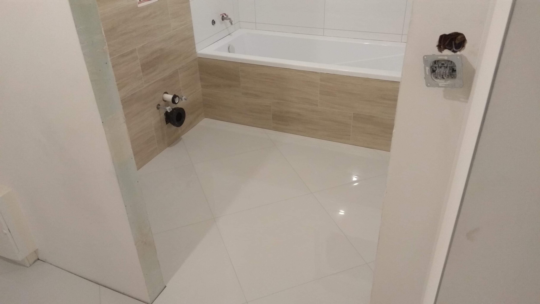 Białe płytki podłogowe w łazience z wanną obudowaną płytkami imitującymi drewno; widoczne odpływy i niedokończone gniazdko elektryczne.