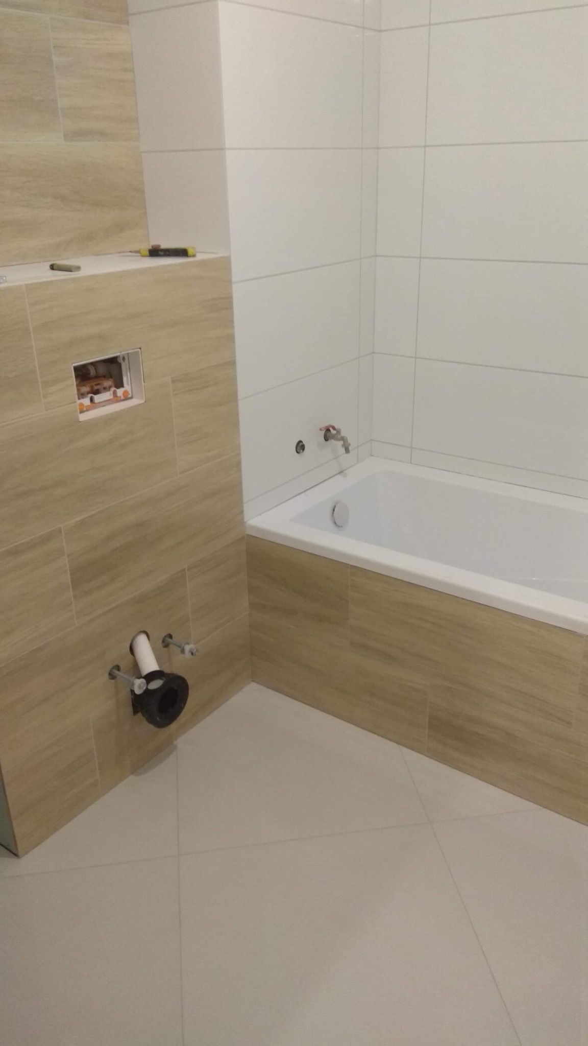 Niedokończona łazienka z beżowymi i białymi płytkami, widoczne rury odpływowe i otwór na instalację elektryczną, narzędzia leżą na półce.