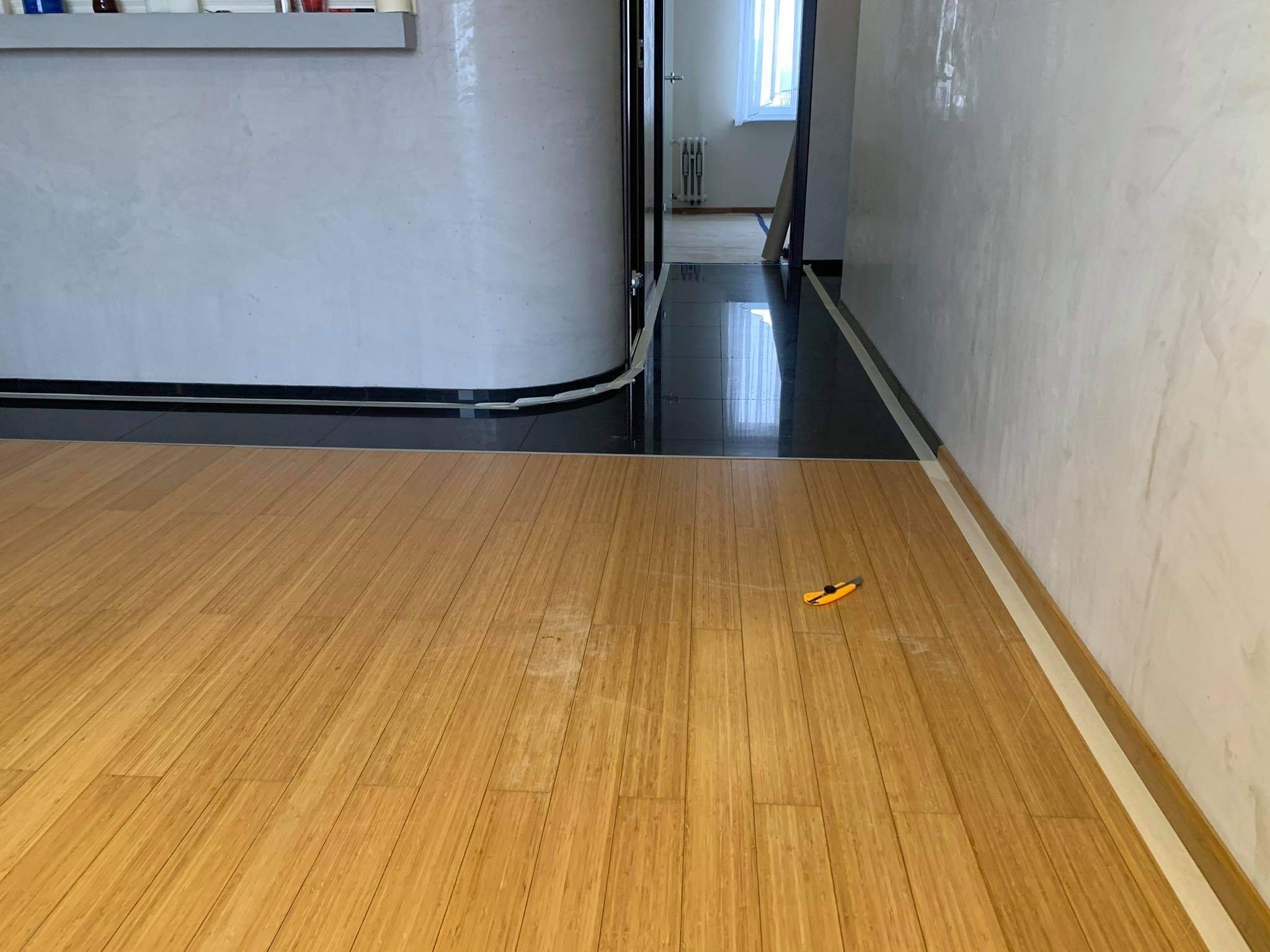 Jasne panele podłogowe z bambusa, z widocznym nożykiem do cięcia paneli i fragmentem ściany z dekoracyjnym tynkiem strukturalnym w tle.