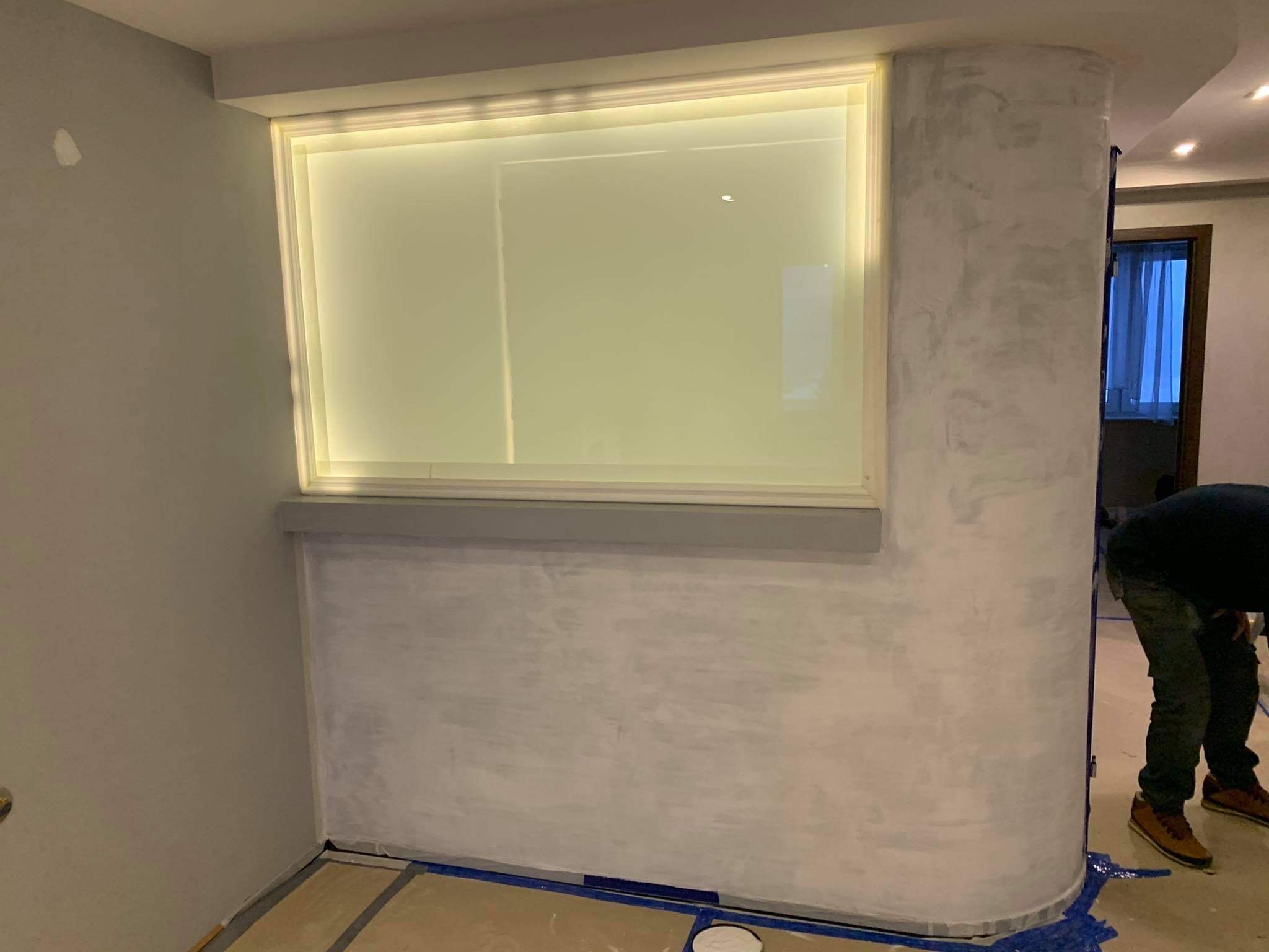 Ściana po nałożeniu szarej farby strukturalnej, z widocznym podświetlanym wnęką i fragmentem pracującego mężczyzny w jeansach.