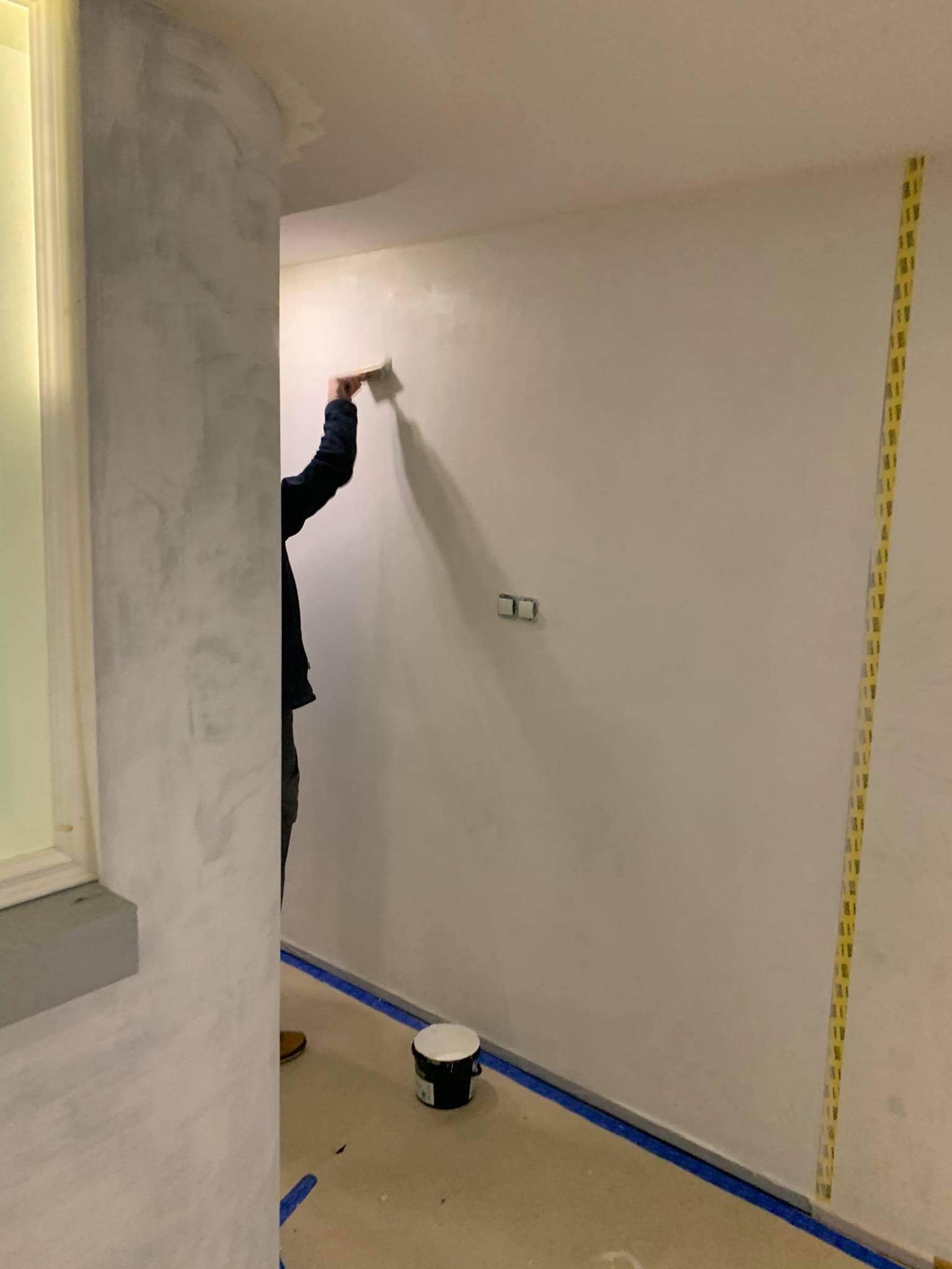 Nakładanie dekoracyjnej farby na ścianę wewnątrz pomieszczenia; widoczny pracownik z narzędziem, wiadro z farbą, zabezpieczone krawędzie taśmą malarską.