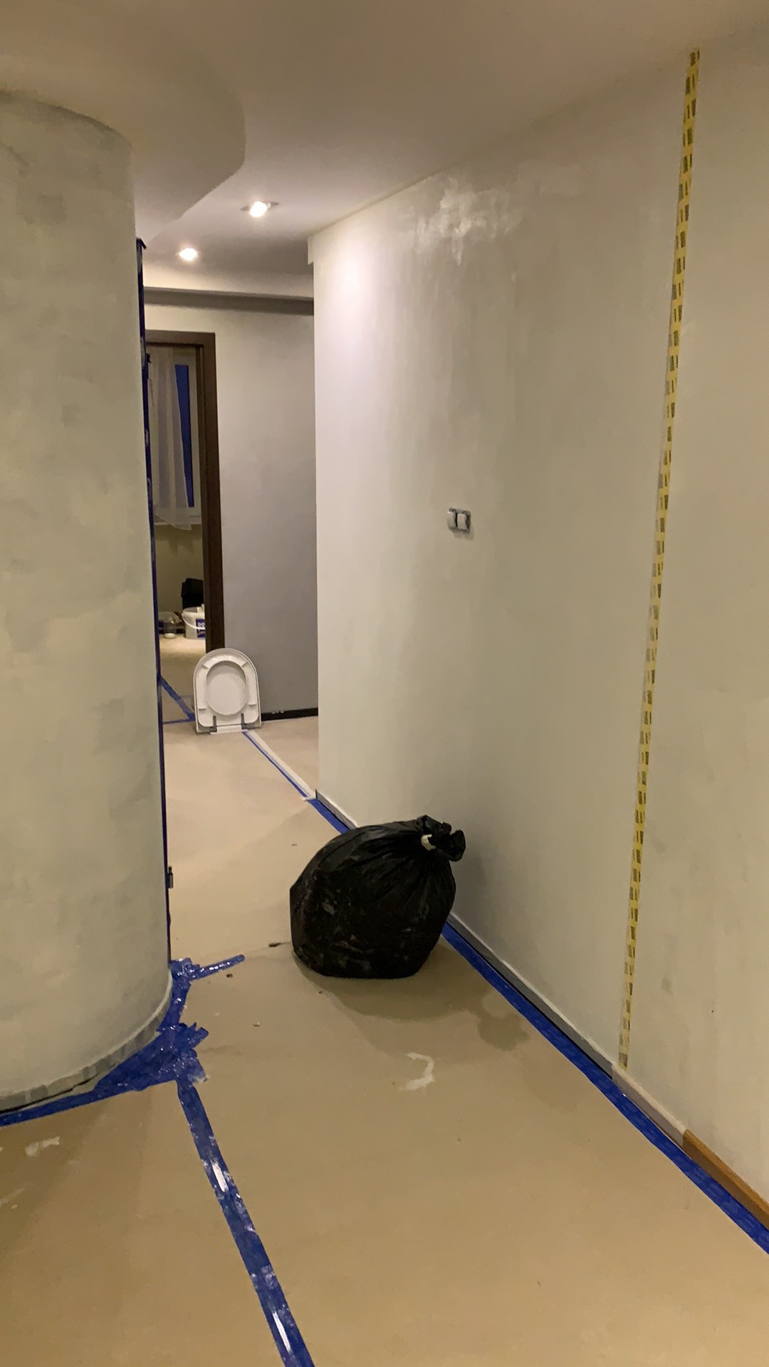 Wnętrze w trakcie prac wykończeniowych, ściany pomalowane na jasny odcień, podłoga zabezpieczona taśmą malarską, widoczny worek na śmieci i demontowana toaleta w tle.