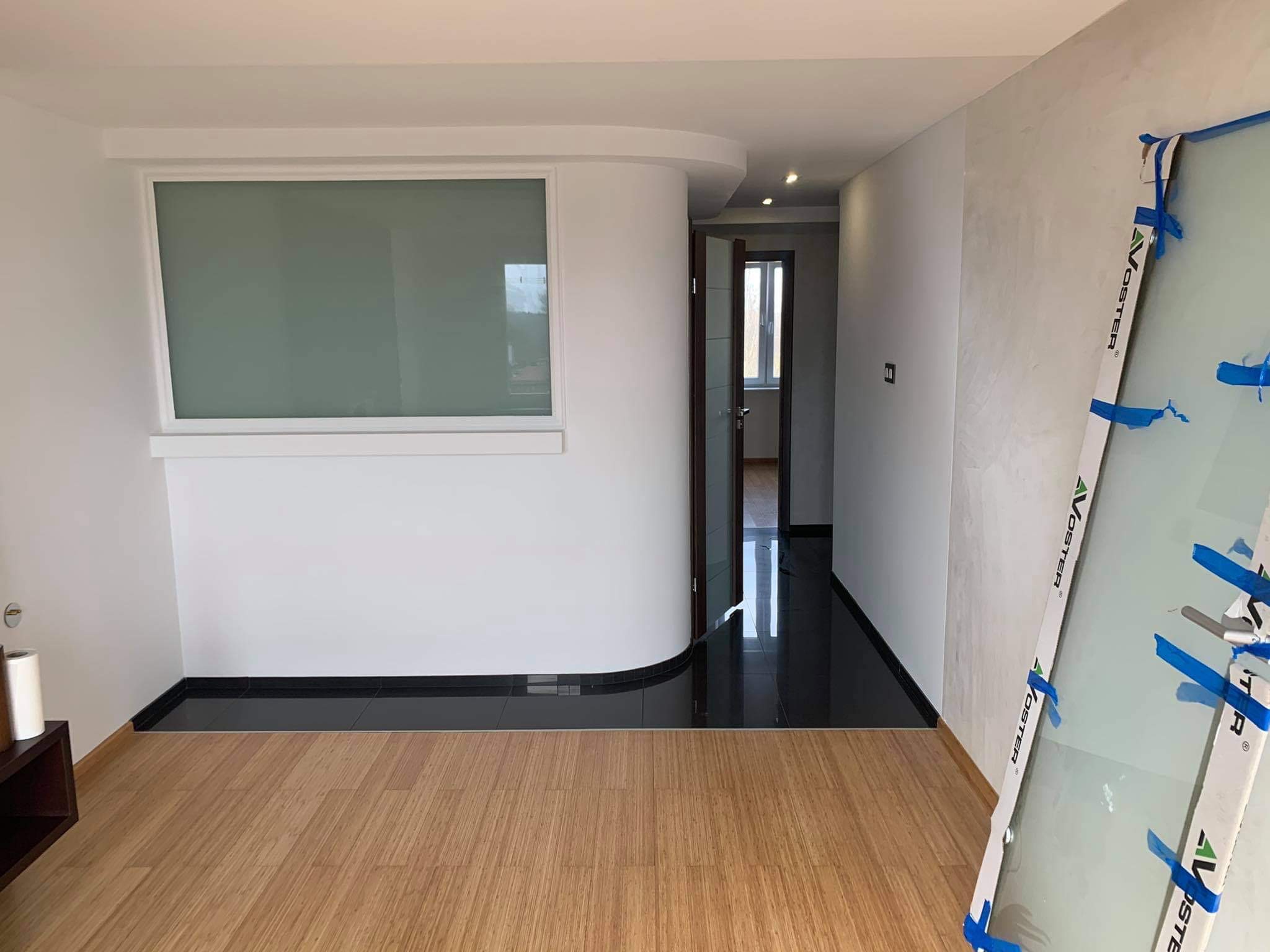 Nowoczesne wnętrze po remoncie: jasne ściany, panele podłogowe w odcieniach drewna, czarne płytki podłogowe przy ścianie, okno z matową szybą i drzwi w głębi, zabezpieczone szkło opierające się...