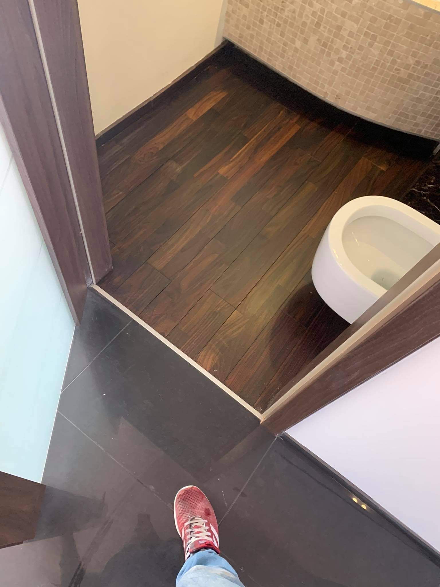Ciemna drewniana podłoga w łazience z mozaikową ścianą, widok z góry na białą toaletę i fragment buta osoby robiącej zdjęcie.