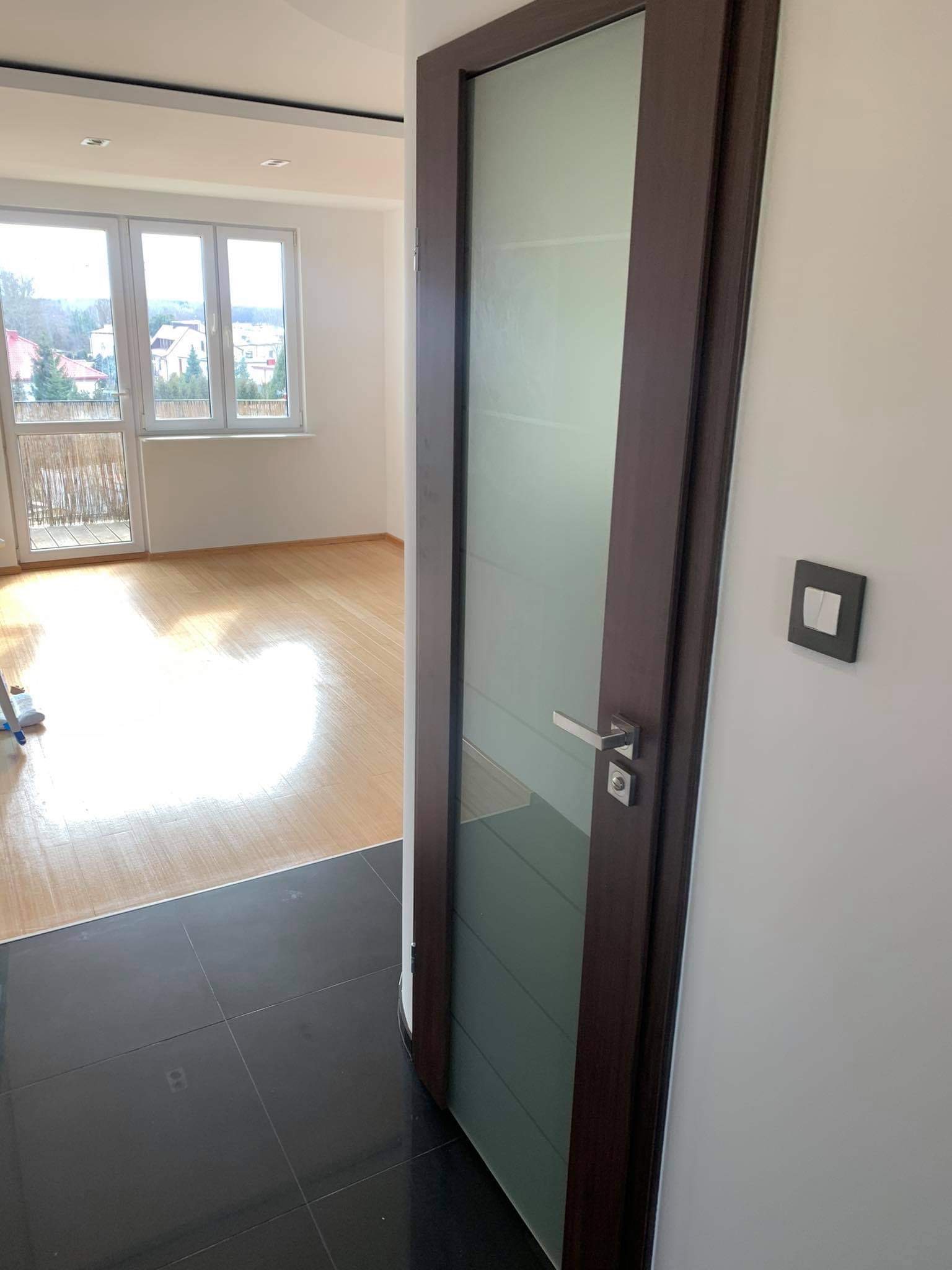 Widok z korytarza na pokój z jasną podłogą i oknem balkonowym, oddzielony ciemnymi płytkami podłogowymi i częściowo otwartymi, przeszklonymi drzwiami z ciemną ramą.