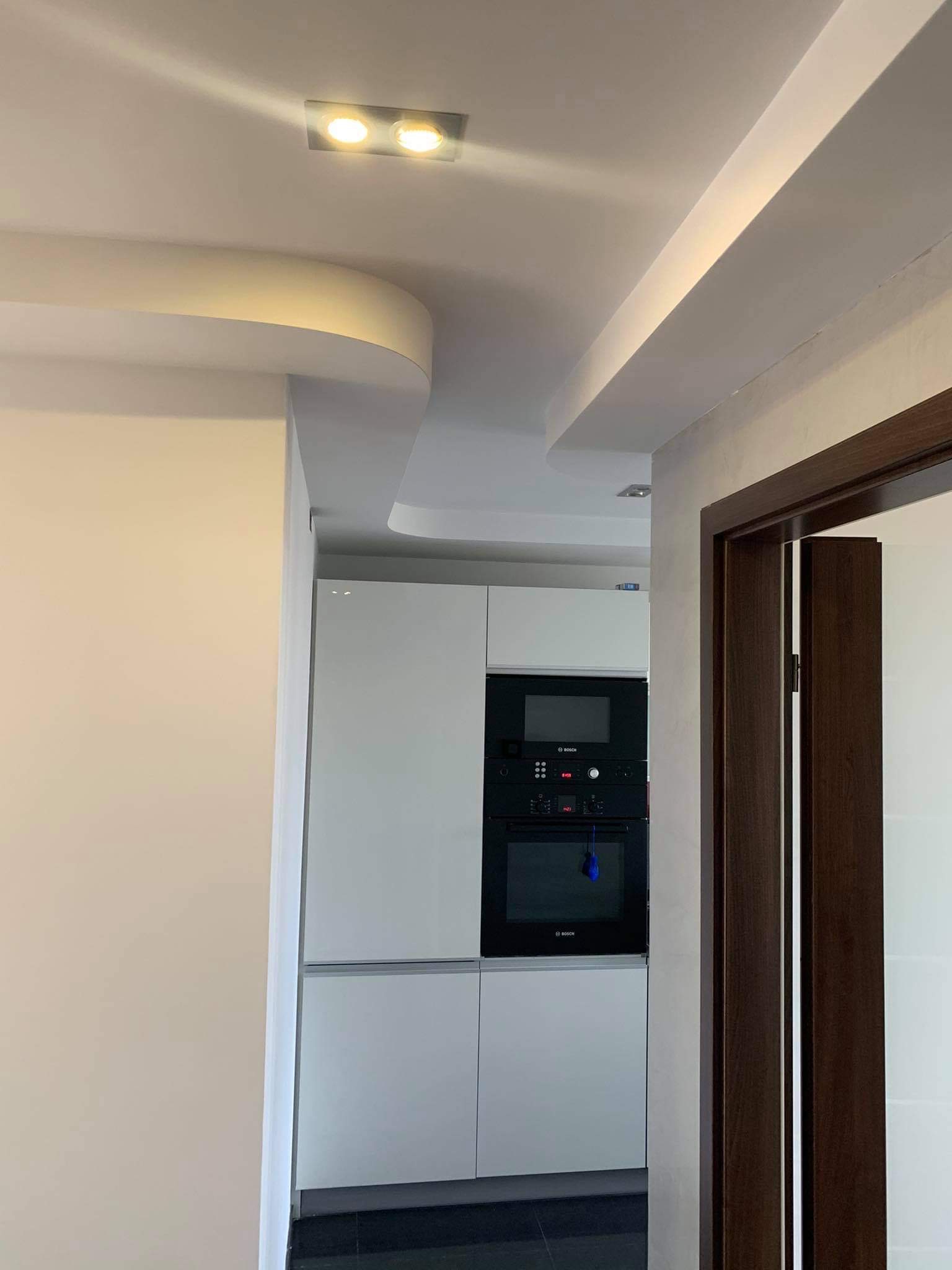 Nowoczesna kuchnia z białymi szafkami i czarnym piekarnikiem Bosch, widoczna przez otwarte drzwi z ciemnobrązową ościeżnicą i dekoracyjnym, podwieszanym sufitem z oświetleniem LED.