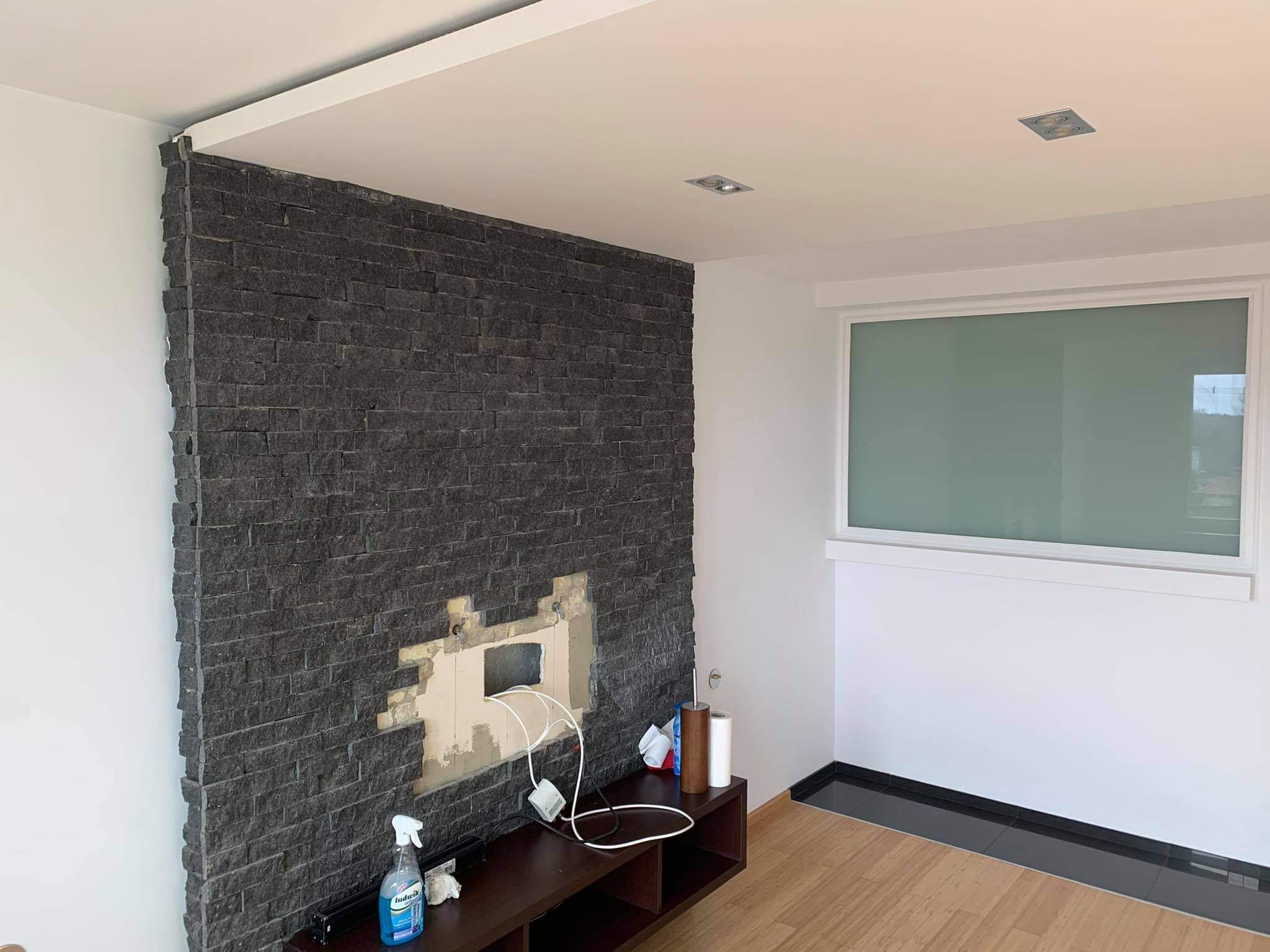 Ściana z dekoracyjnej, czarnej cegły z widocznym miejscem po demontażu elementów, butelka środka czyszczącego na szafce RTV, jasne panele podłogowe i białe ściany.