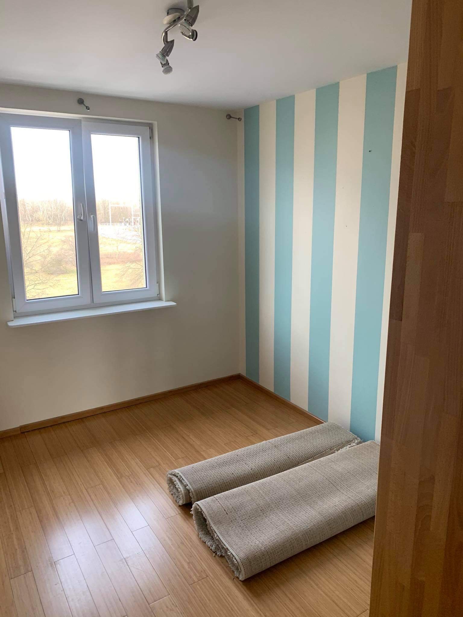 Pusty pokój z drewnianą podłogą, ścianą w biało-błękitne pasy i zwiniętymi dywanikami, przygotowany do prac wykończeniowych.