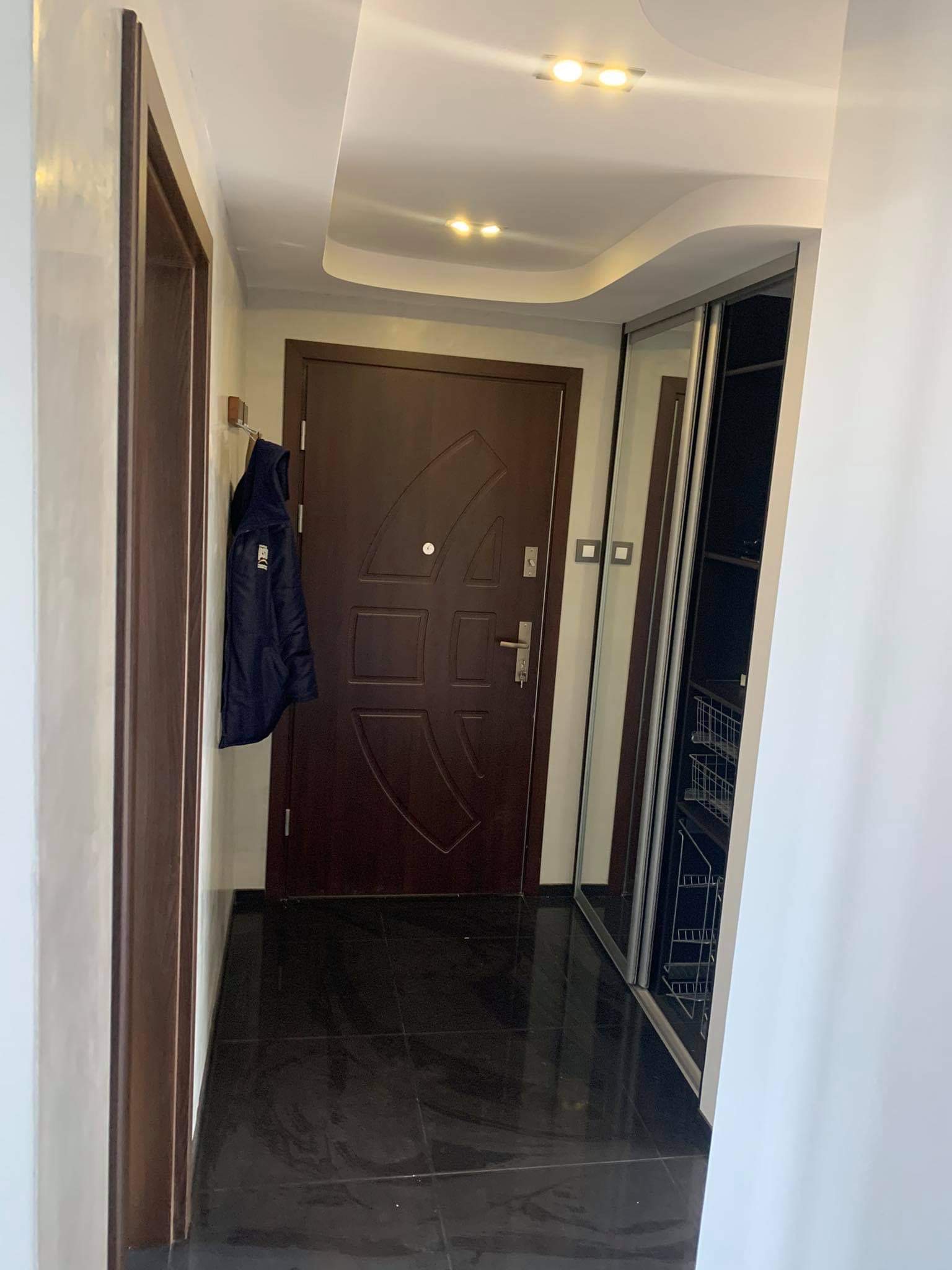 Przedpokój z ciemną podłogą, brązowymi drzwiami wejściowymi z metalowymi elementami, przesuwną szafą z lustrami i kurtką wiszącą na wieszaku.