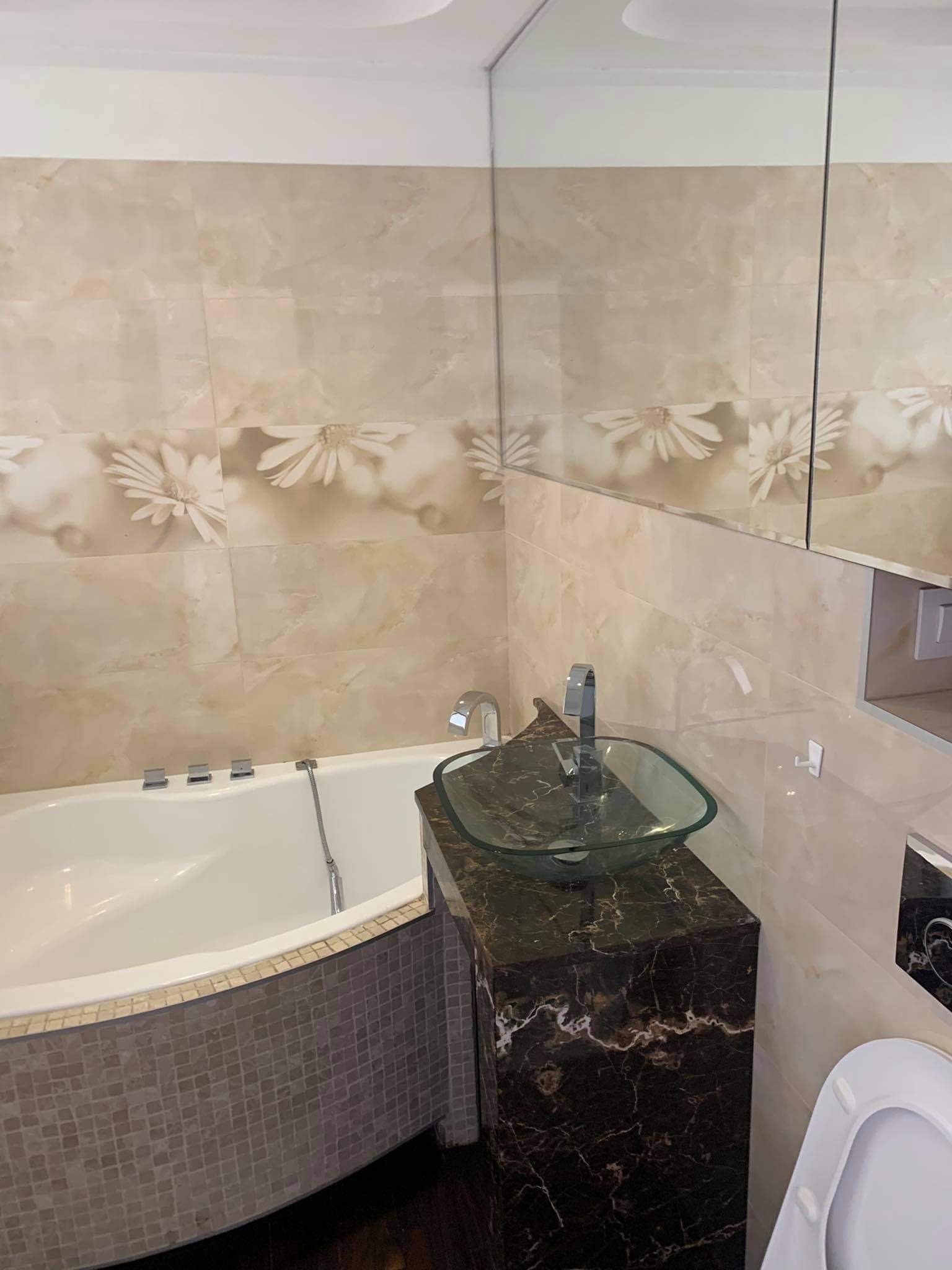 Nowoczesna łazienka z wanną obudowaną mozaiką, szklaną umywalką na postumencie z czarnego marmuru i płytkami ściennymi w odcieniach beżu z motywem kwiatowym.