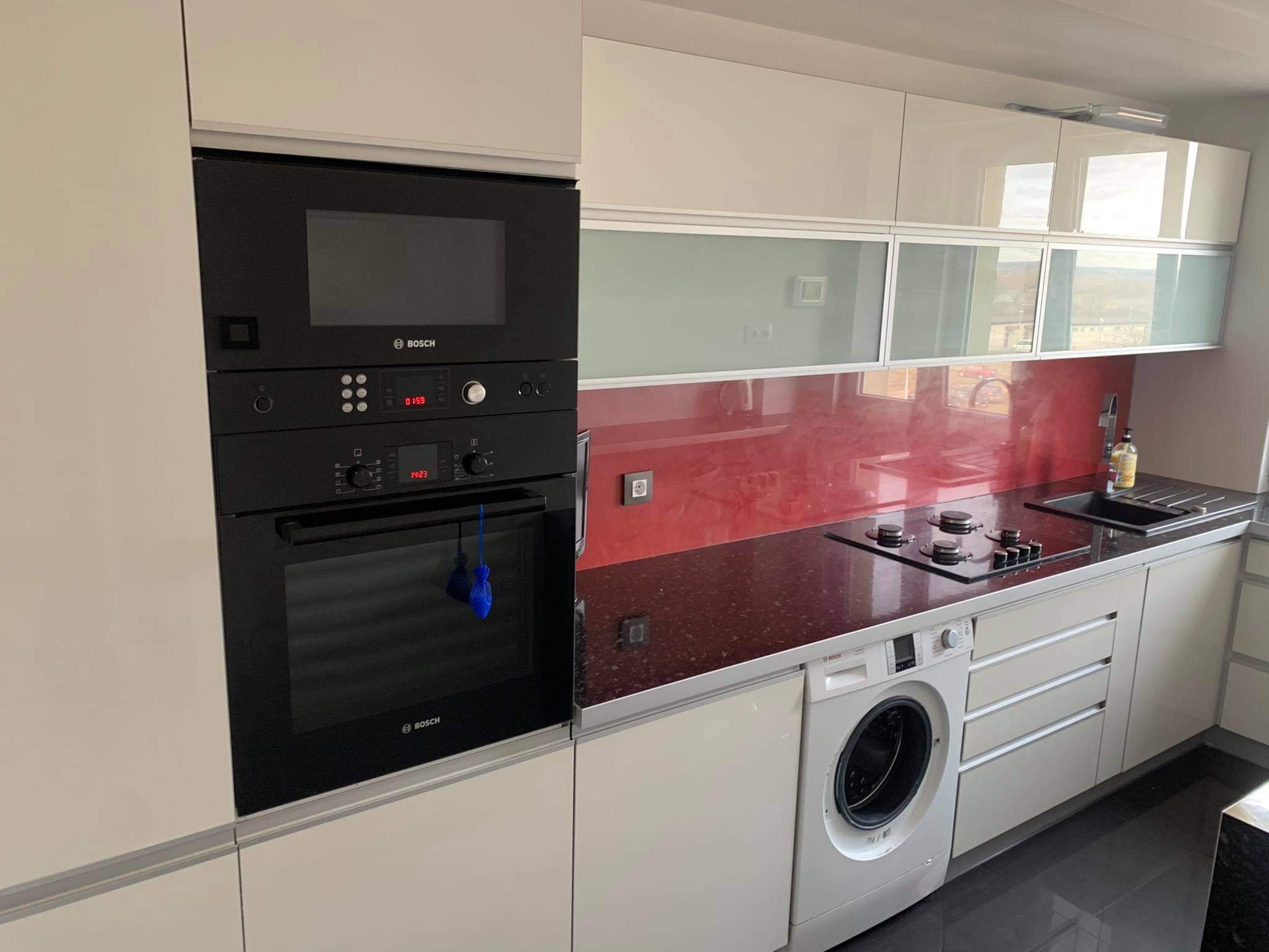 Nowoczesna kuchnia z zabudową w jasnych odcieniach, z wbudowanym sprzętem AGD marki Bosch, ciemnym blatem i czerwonym panelem szklanym nad blatem, oraz pralką zintegrowaną z zabudową.