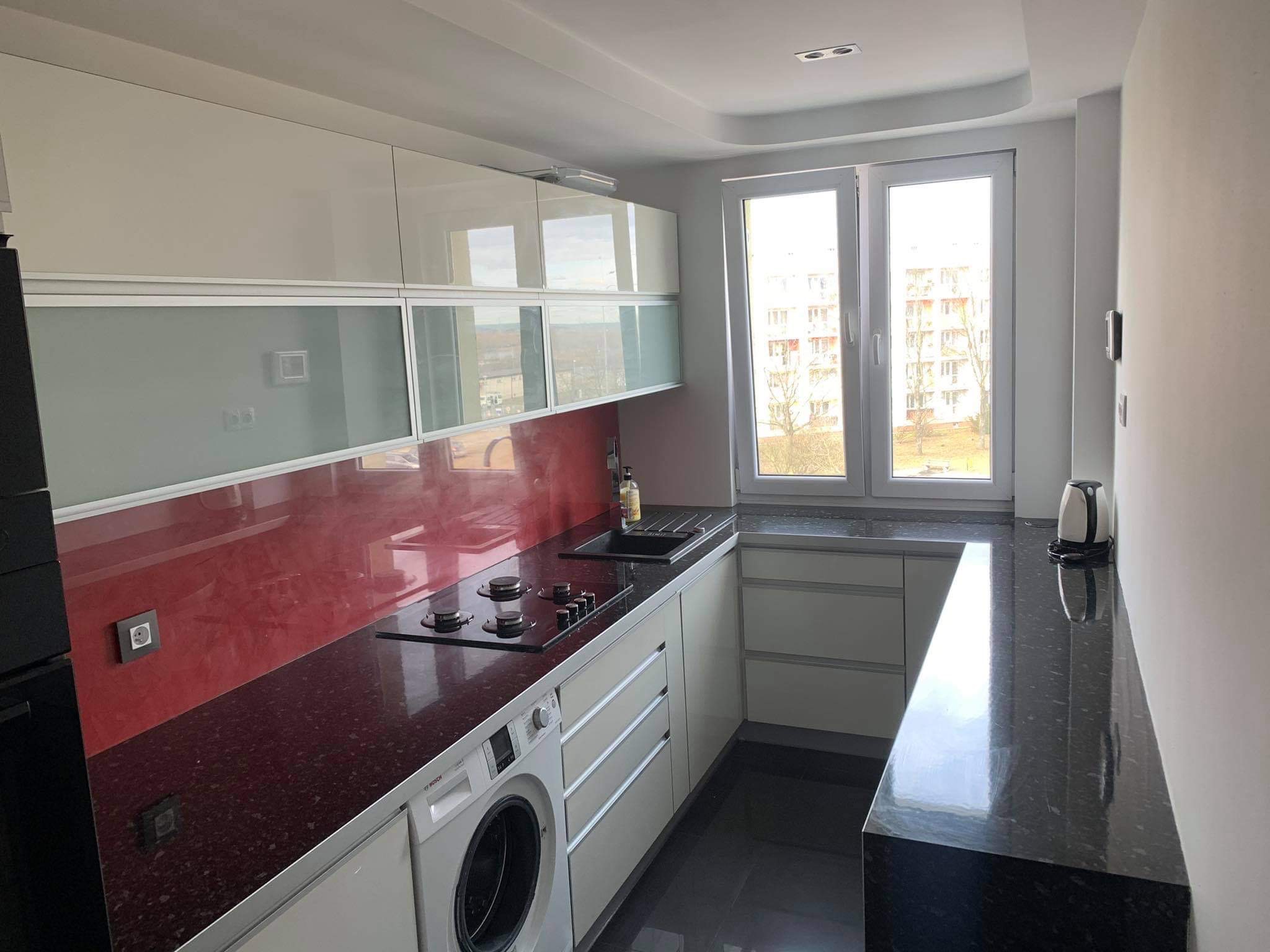 Wnętrze nowoczesnej kuchni z białymi szafkami, czerwonym panelem nad blatem z czarnego granitu, zintegrowaną pralką Bosch i widokiem z okna na budynek mieszkalny.