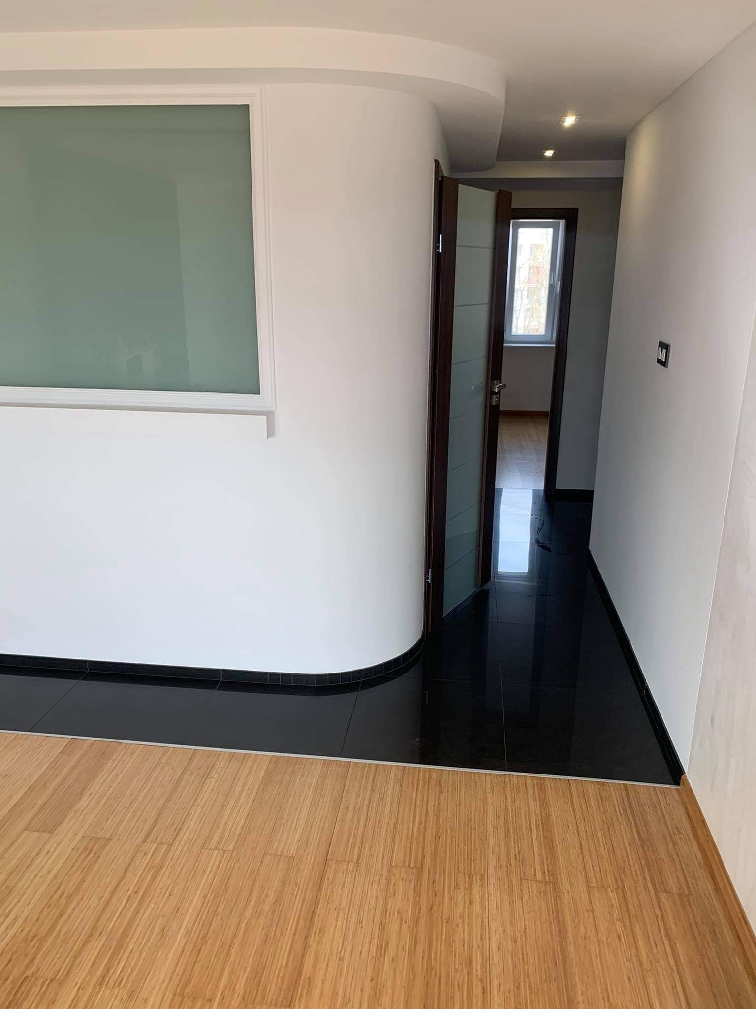 Wnętrze mieszkania po remoncie: połączenie jasnej drewnianej podłogi, czarnej błyszczącej posadzki i białych ścian z zaokrąglonym narożnikiem, widok na otwarte drzwi do kolejnego pomieszczenia.