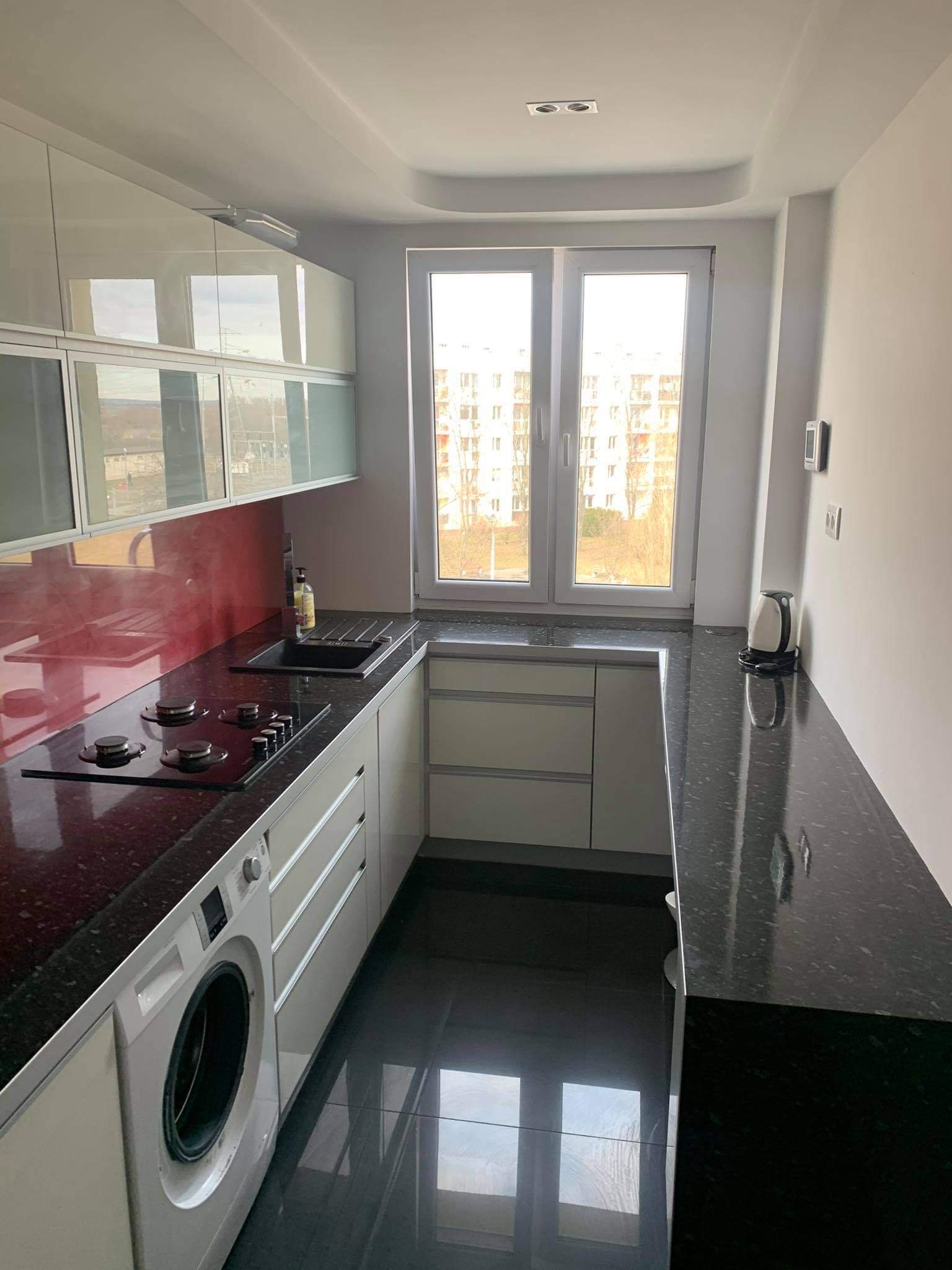 Nowoczesna, jasna kuchnia z czarnym blatem i błyszczącą podłogą, z pralką w zabudowie pod blatem, widok na okno z budynkami w tle.