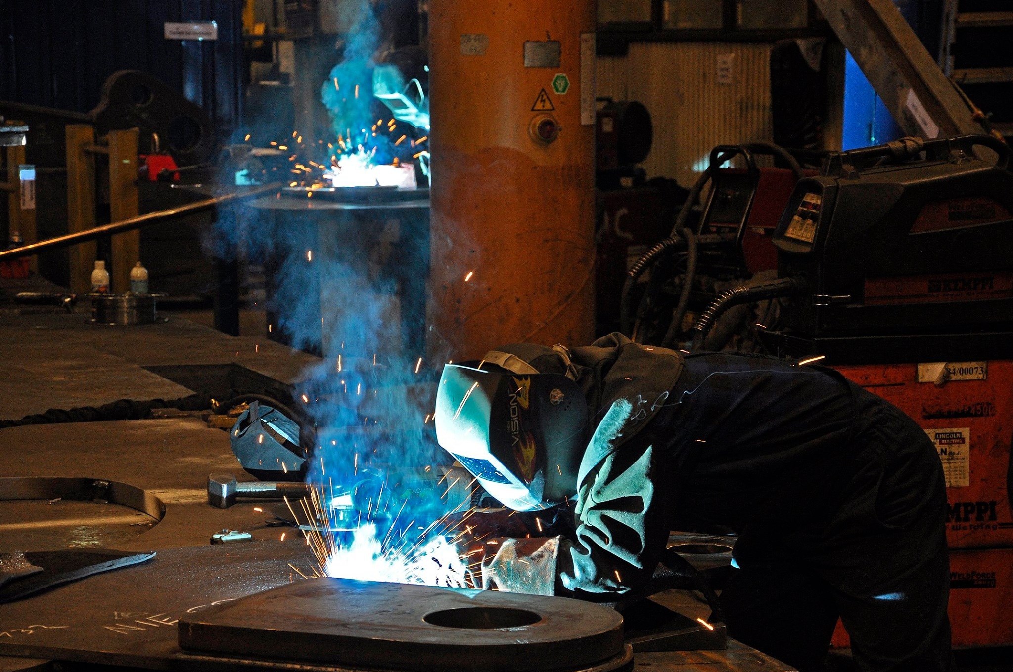 Spawacz w masce ochronnej spawający element konstrukcyjny, iskry i dym w warsztacie z pomarańczowym cylindrem i spawarką w tle.