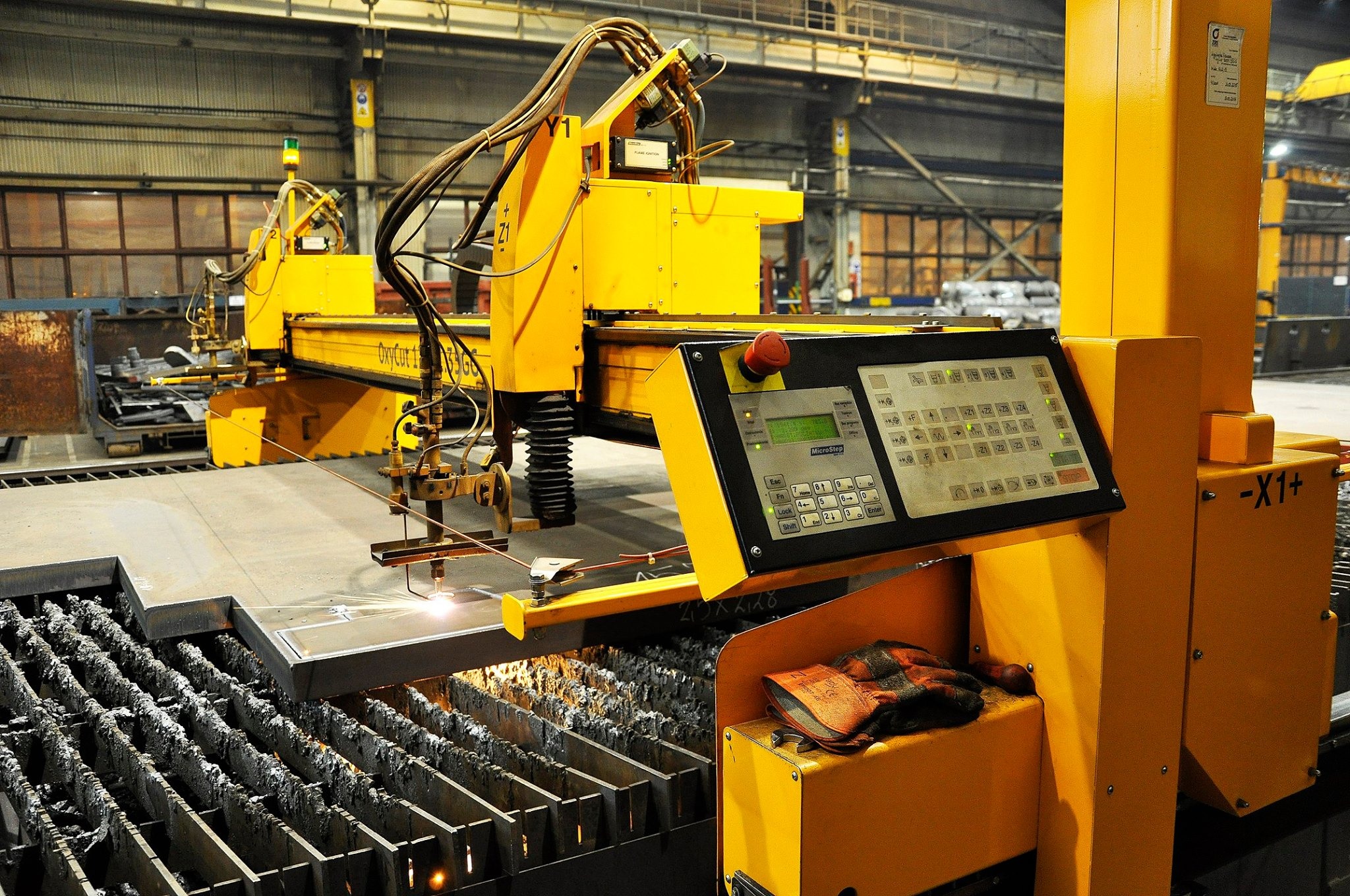 Żółta maszyna CNC do cięcia metalu w hali produkcyjnej, iskry podczas cięcia, panel sterowania z klawiaturą i ekranem, na maszynie leżą rękawice robocze.